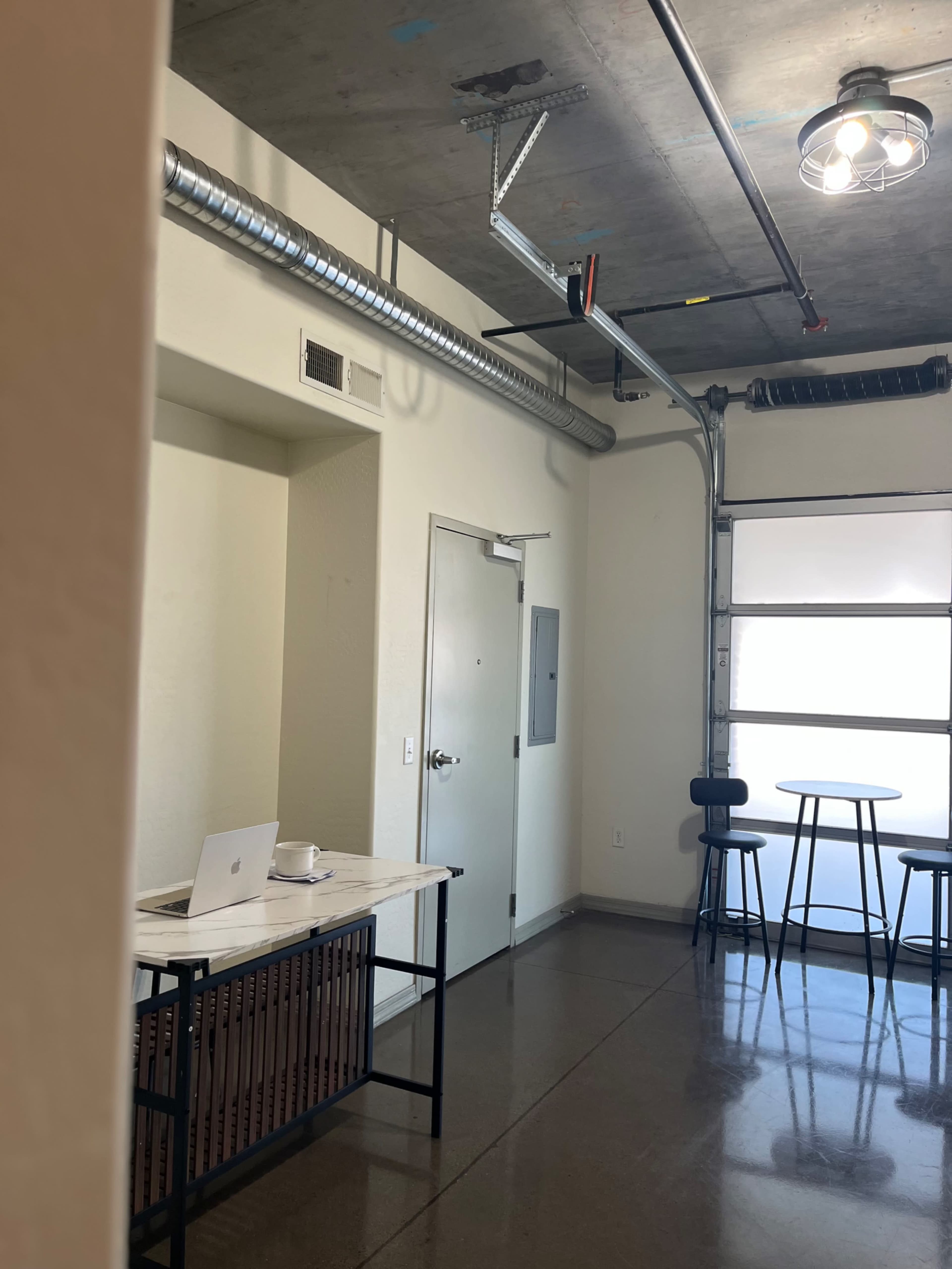 The image shows a modern, minimalist workspace with a table and bar stools, a garage door, and exposed piping on the ceiling.