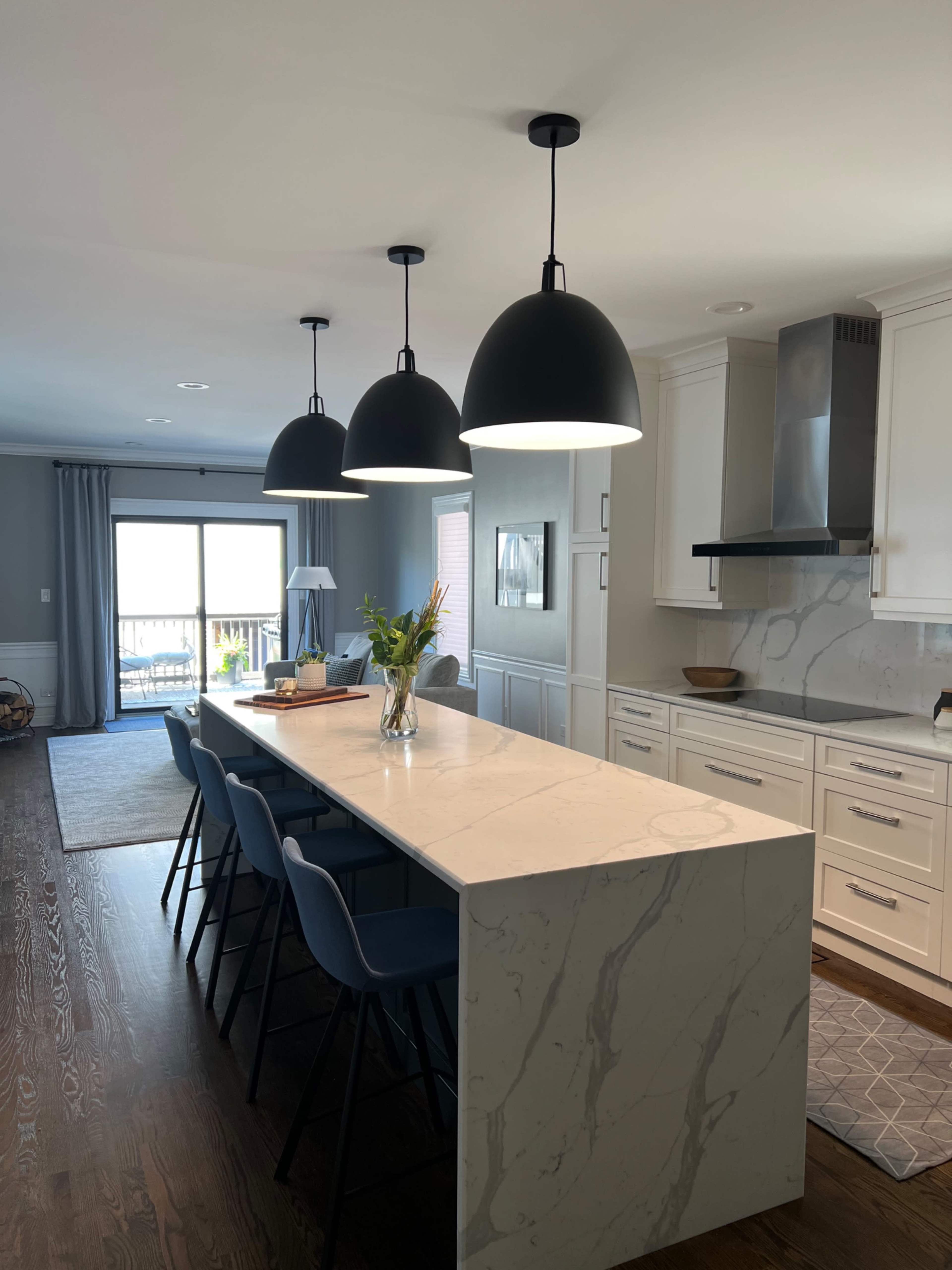 A modern kitchen features a marble island with black pendant lights and blue upholstered bar stools, overlooking a sliding glass door that leads to an outdoor area.