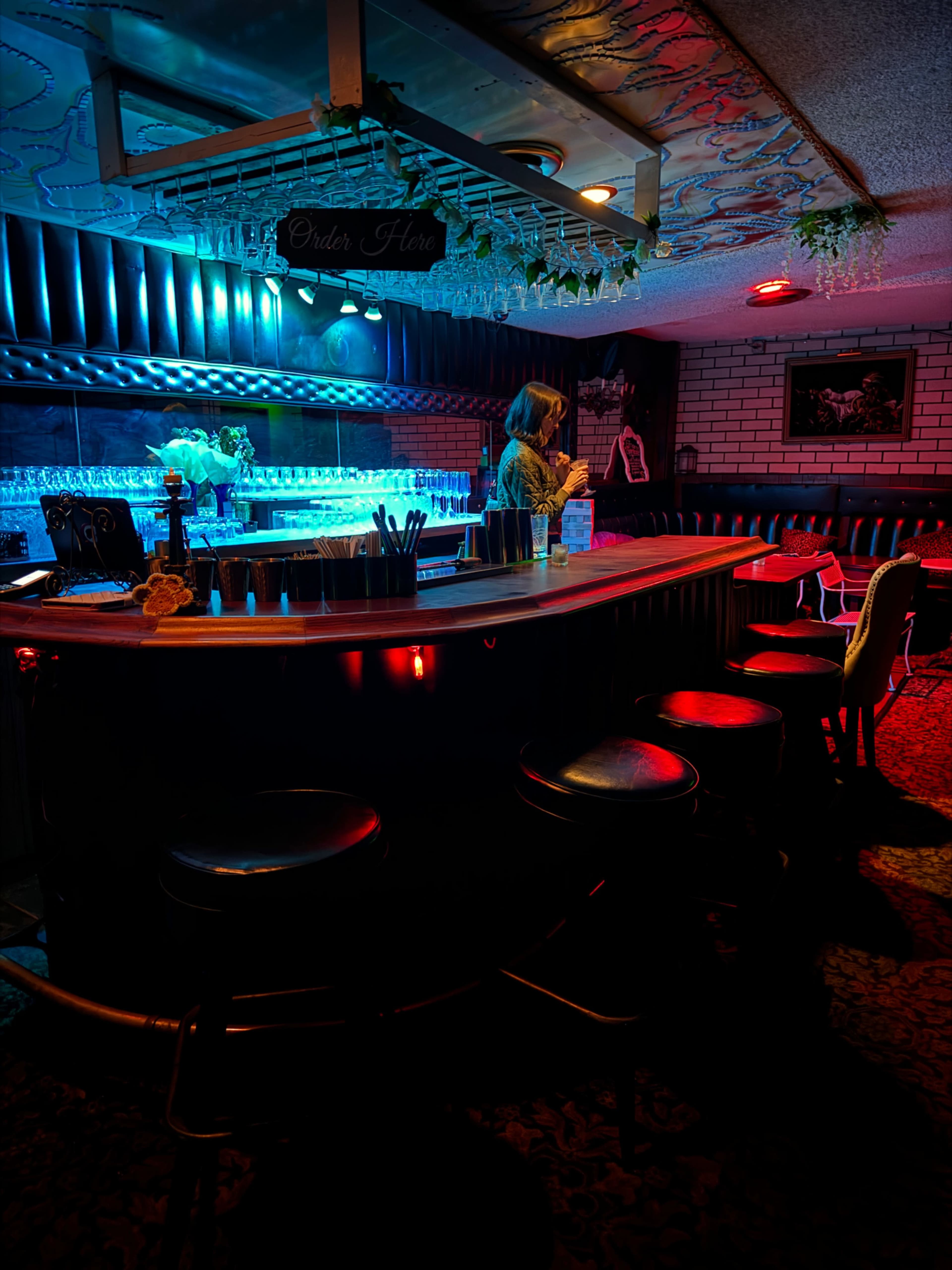 A dimly lit bar features a curved counter with high stools and a bartender preparing drinks under colorful lighting.