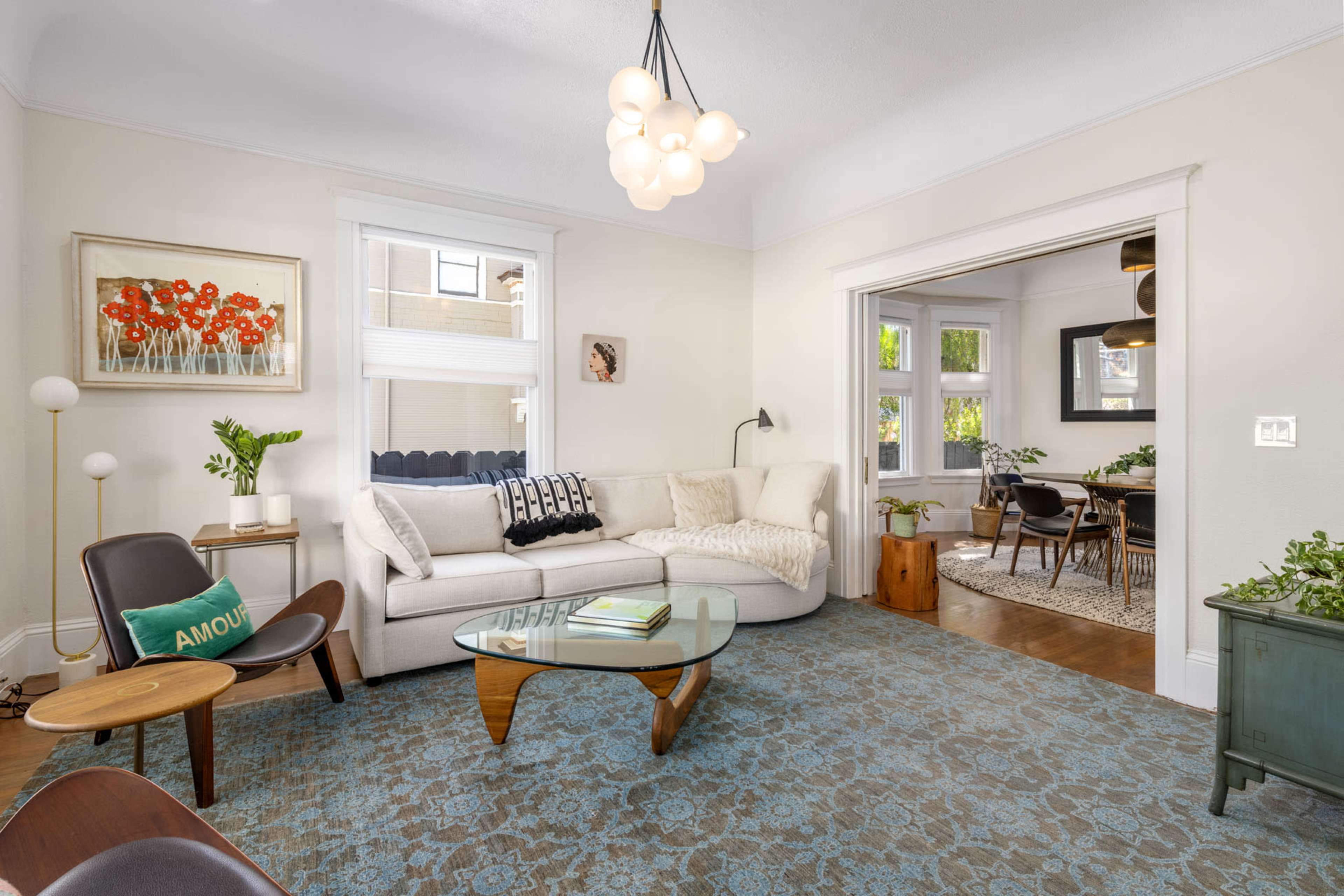 A well-lit living room featuring a white sectional sofa, a glass coffee table, and decorative plants, with an adjoining room visible in the background.