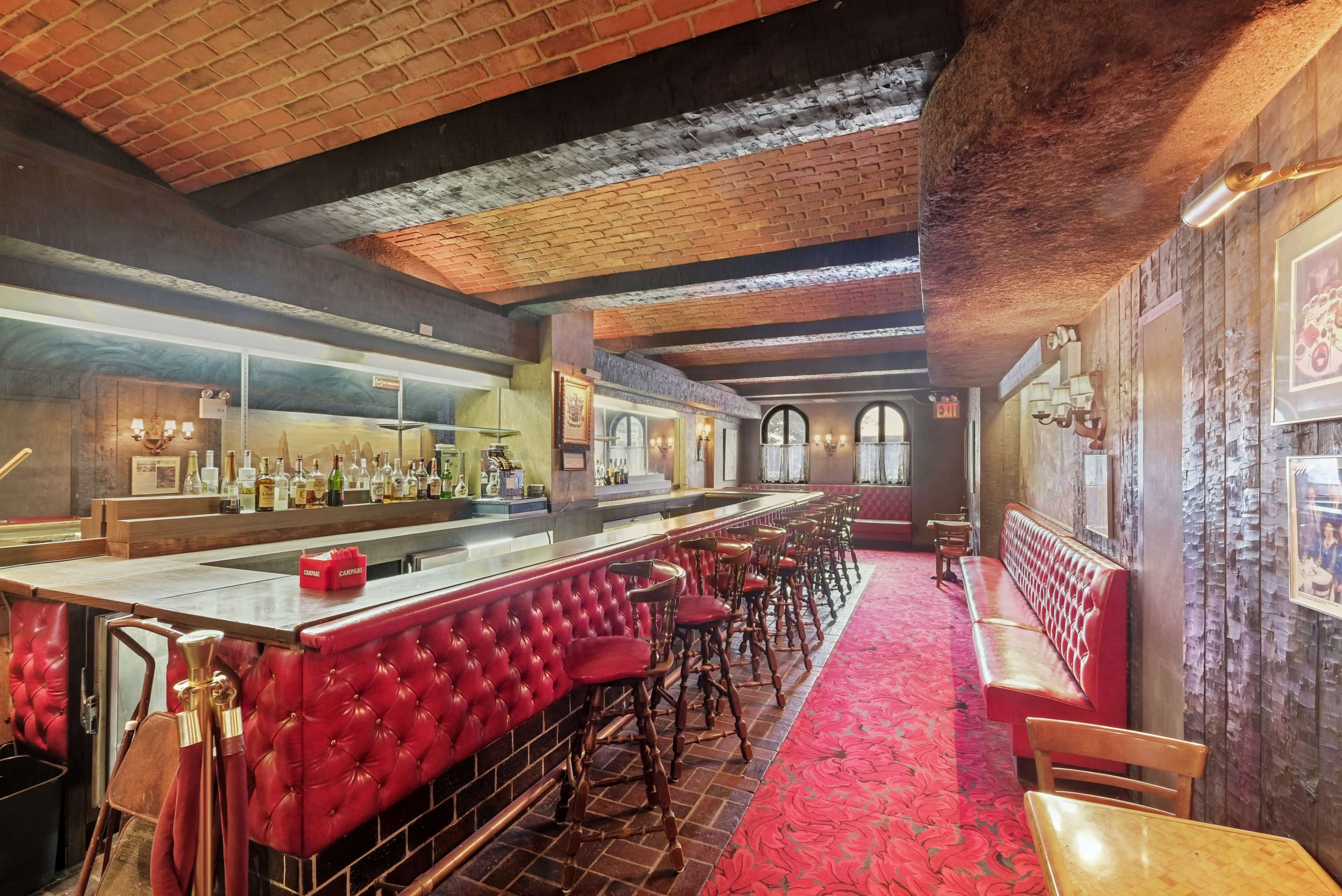 A dimly lit bar with red upholstered seating, a long marble-topped counter, and shelves lined with bottles in the background.