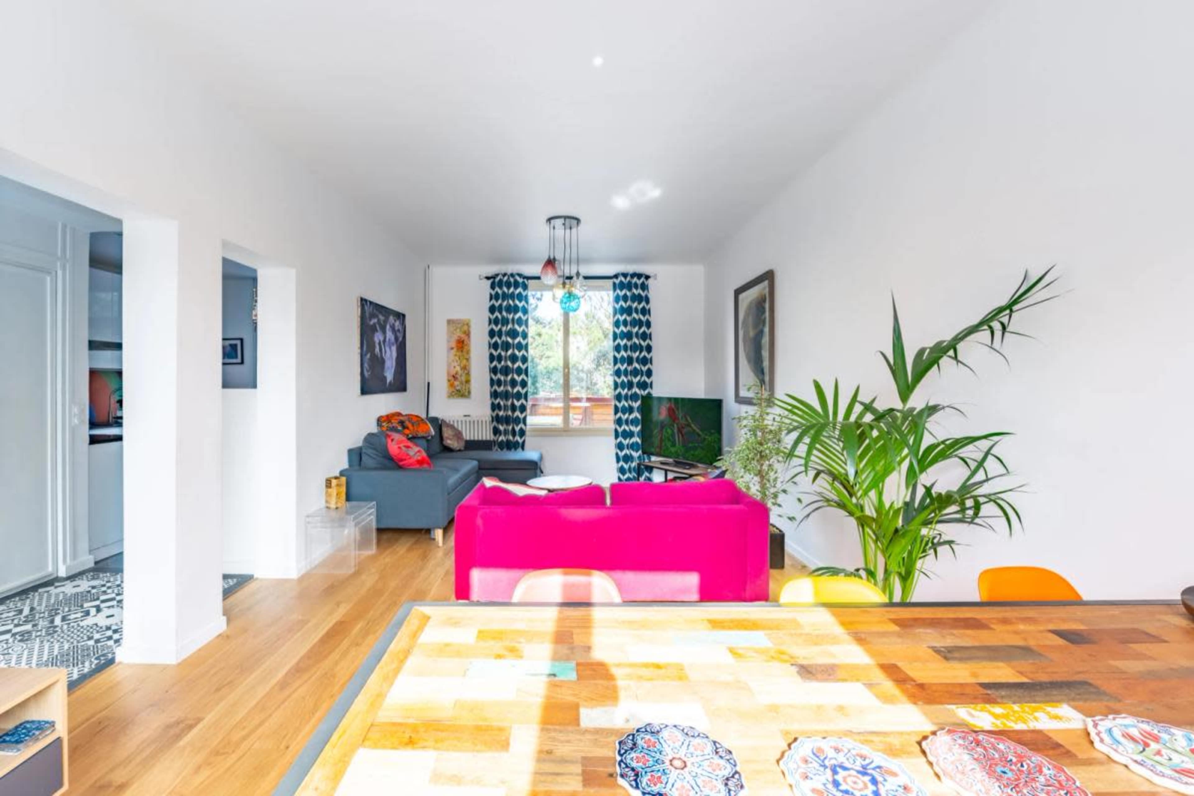 A bright living space with a dining table in the foreground, a gray sofa, and colorful decor, including curtains and cushions.