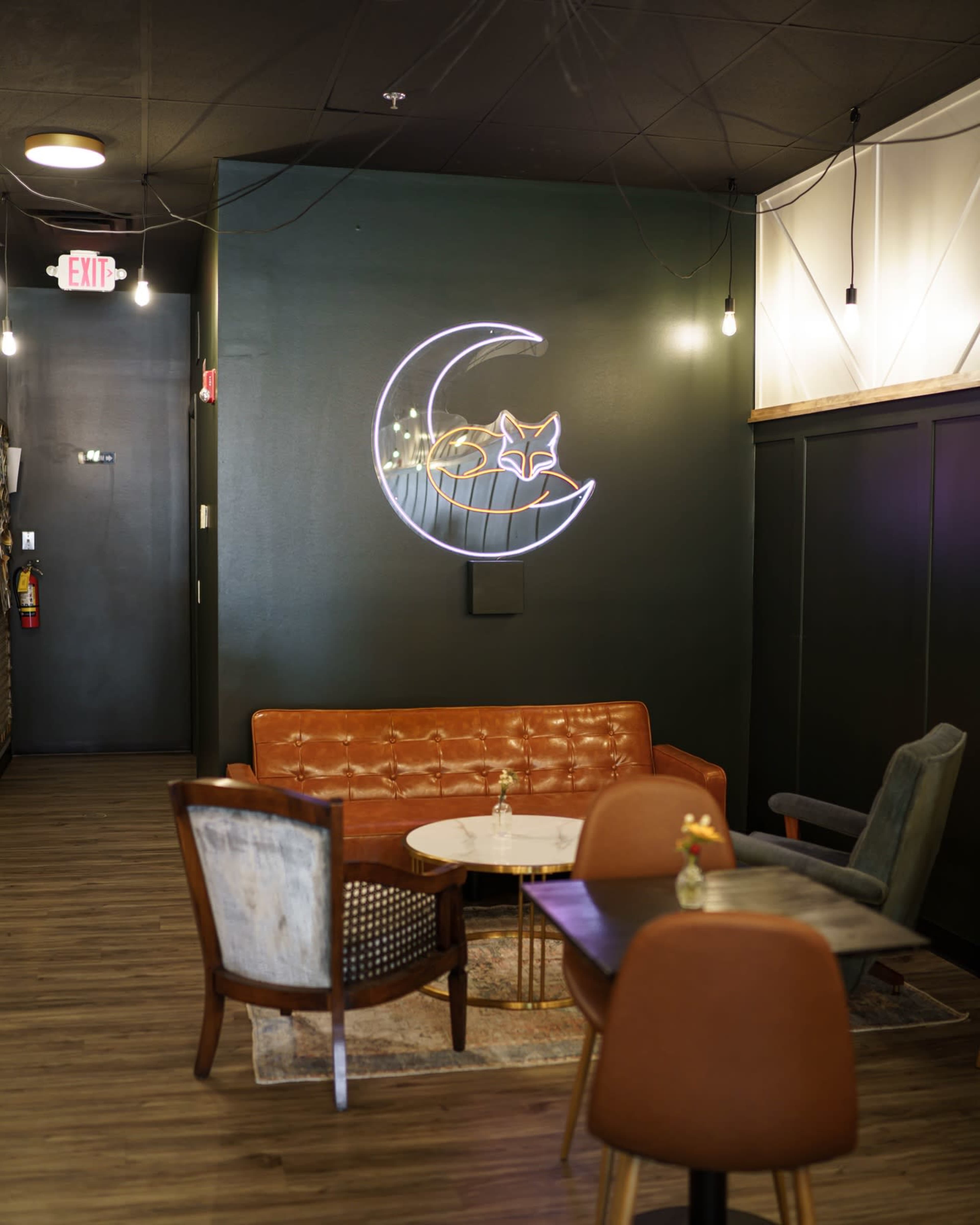 The image shows a cozy lounge area featuring a brown leather sofa, mixed seating options, a round table in the center, and a neon sign of a moon and a fox on the dark green wall.
