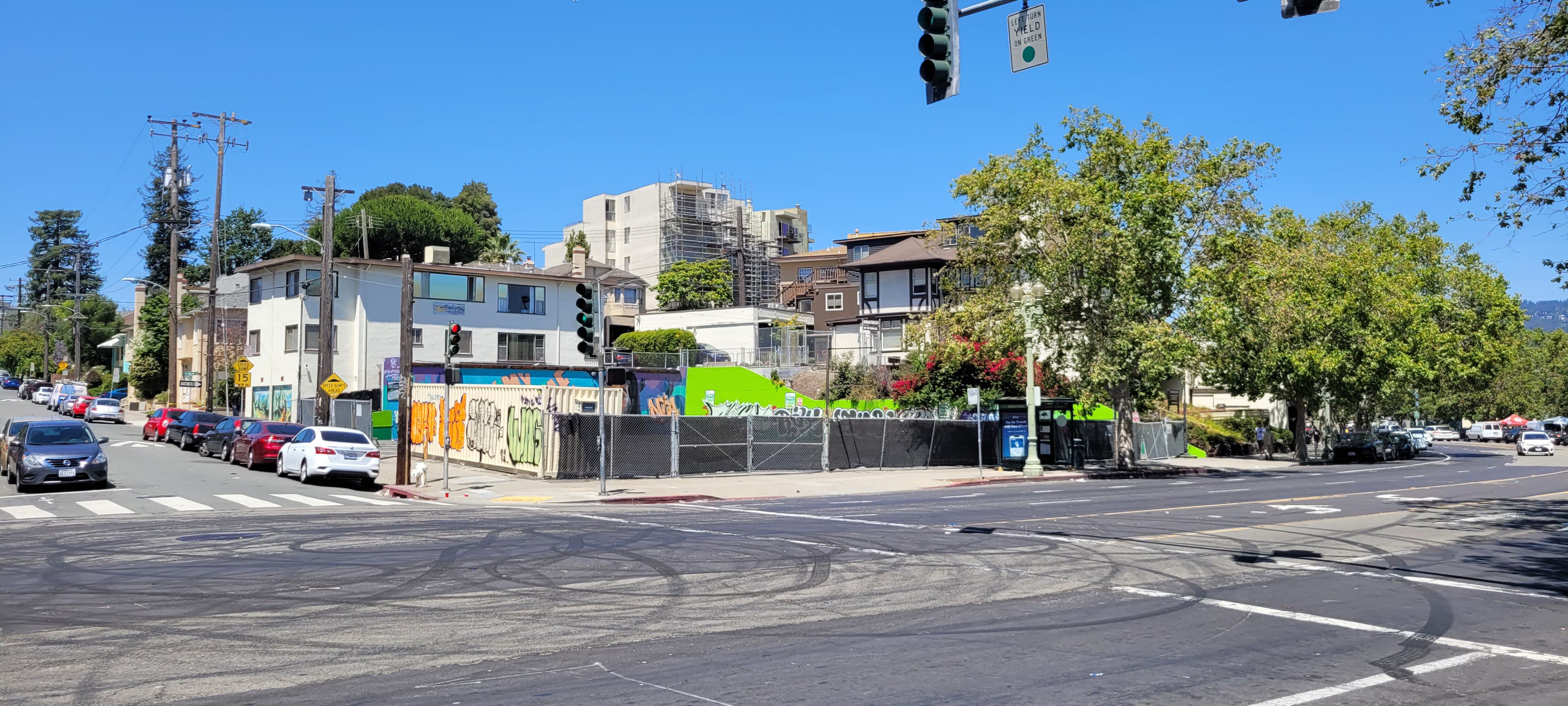 The image shows an urban intersection with a green mural on a fence, surrounded by residential buildings and trees under a clear blue sky.