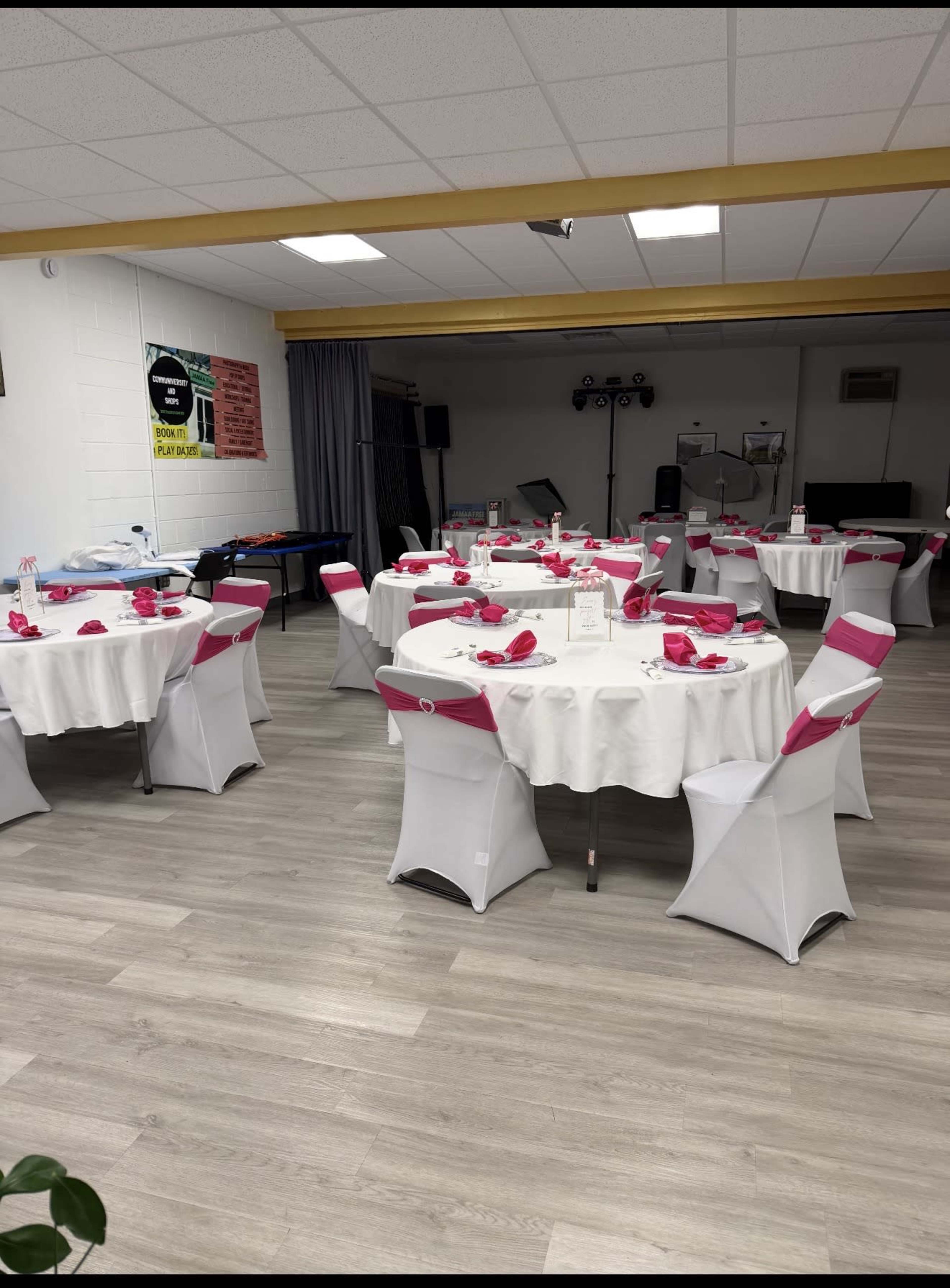 The image shows a banquet hall with several round tables covered in white cloths and adorned with pink napkins, arranged neatly in a spacious, well-lit area.