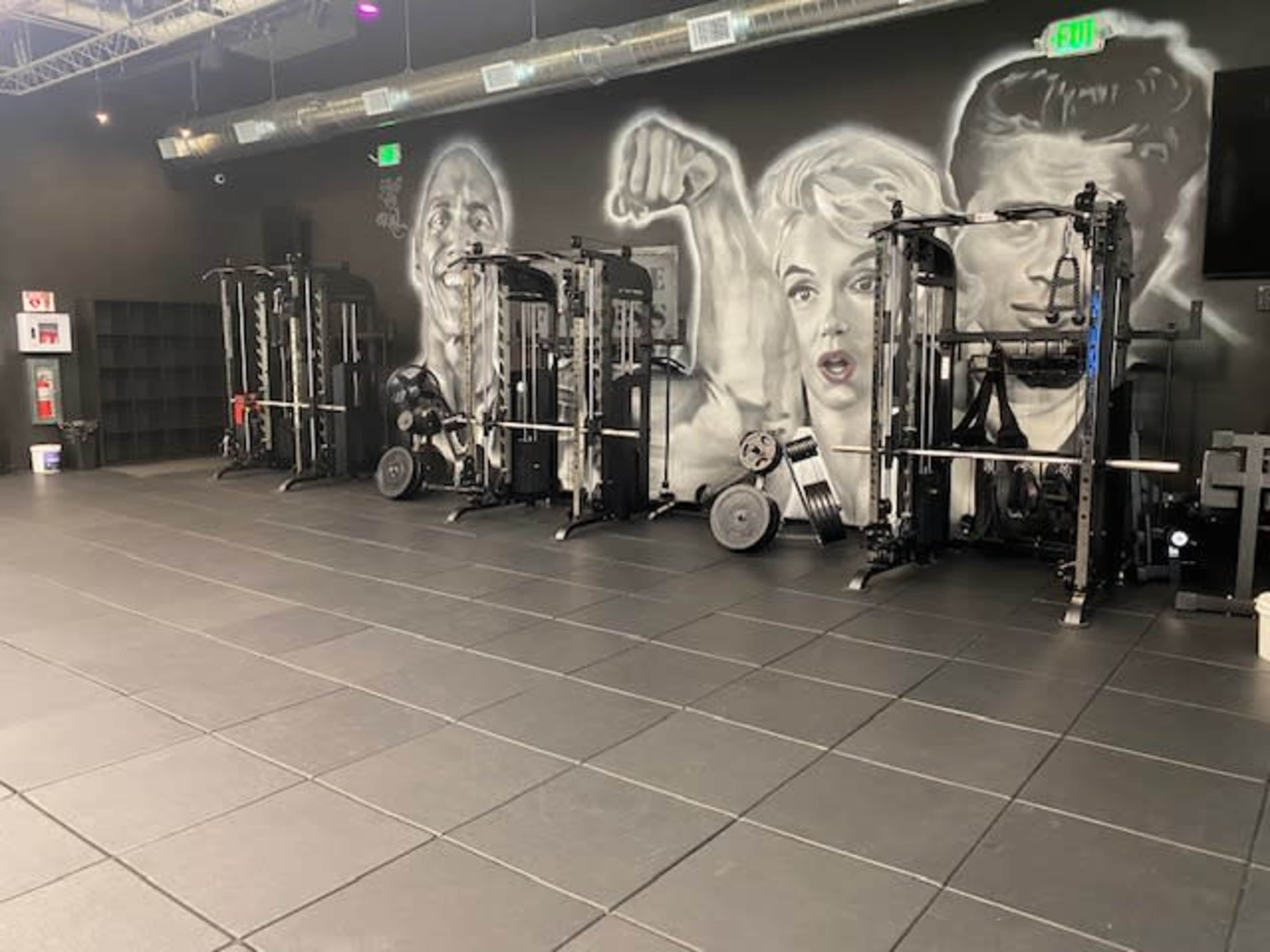 The image shows a gym with multiple weightlifting machines against a wall featuring black-and-white portraits of muscular figures.