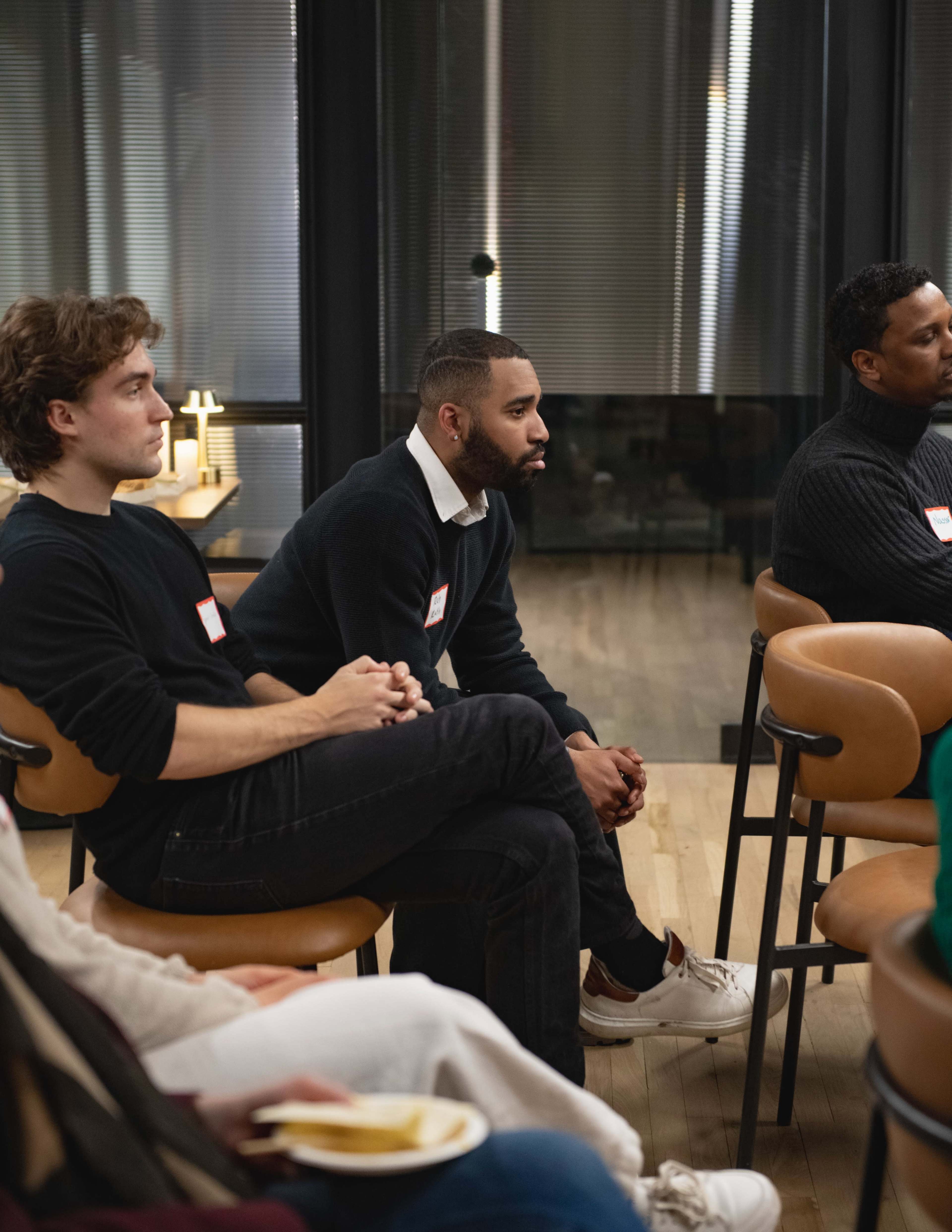A group of people sits in a modern conference room, attentively listening to a speaker, with name tags visible on their clothing.