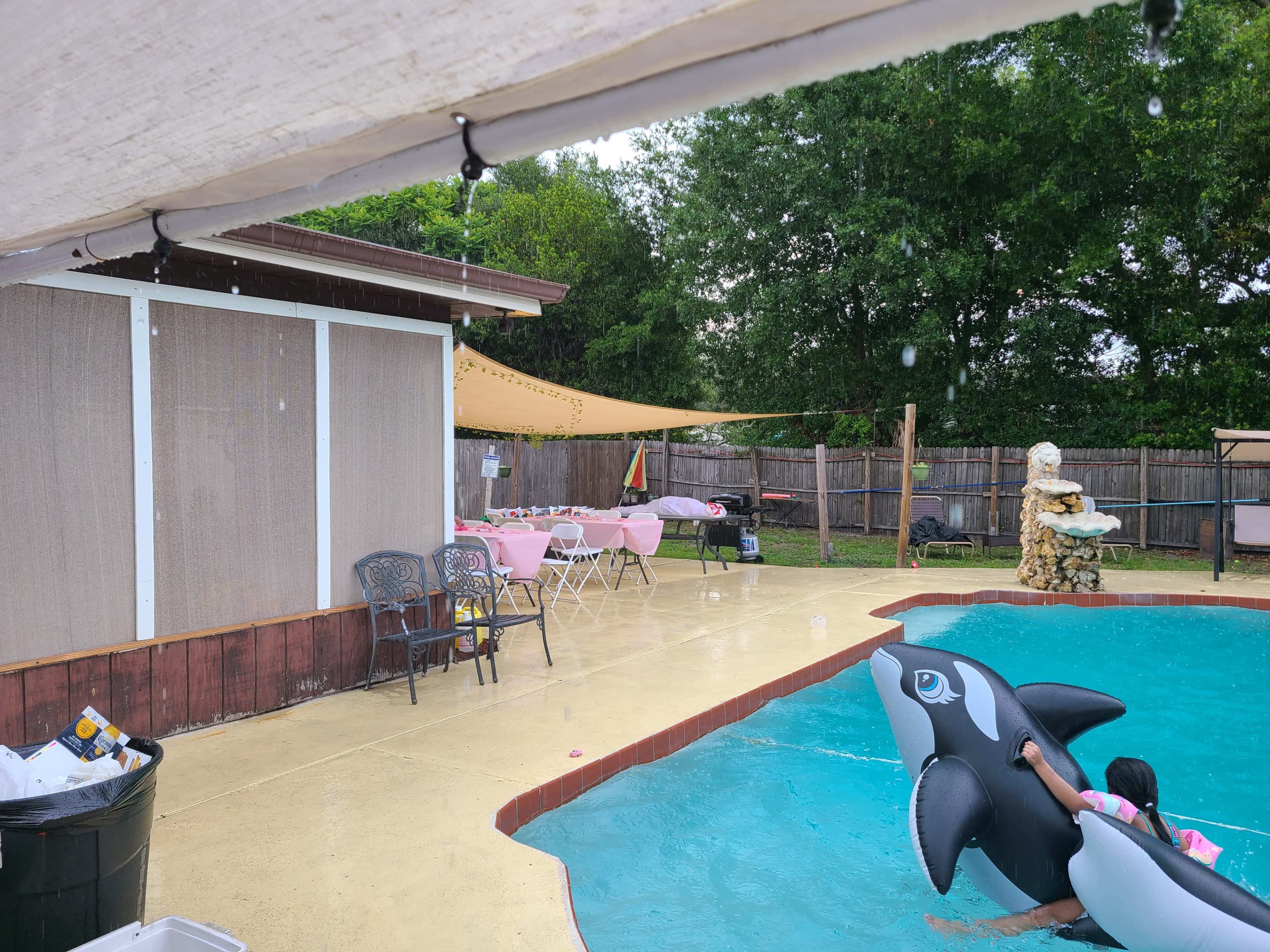 The scene shows a backyard pool area with a person on a floating device, surrounded by tables set up for an event and a covered seating area.