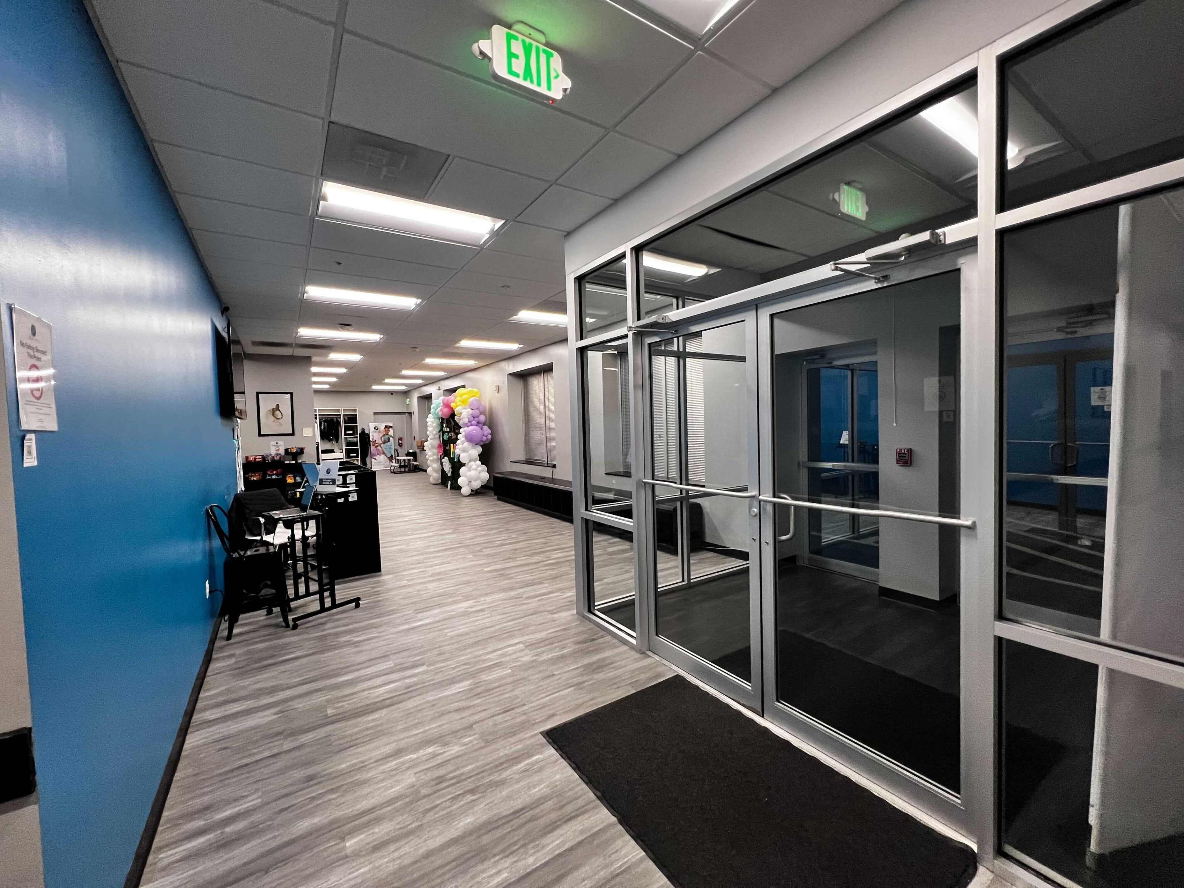 A well-lit hallway leads to a glass entrance, with seating, decorations on the side, and an exit sign above.