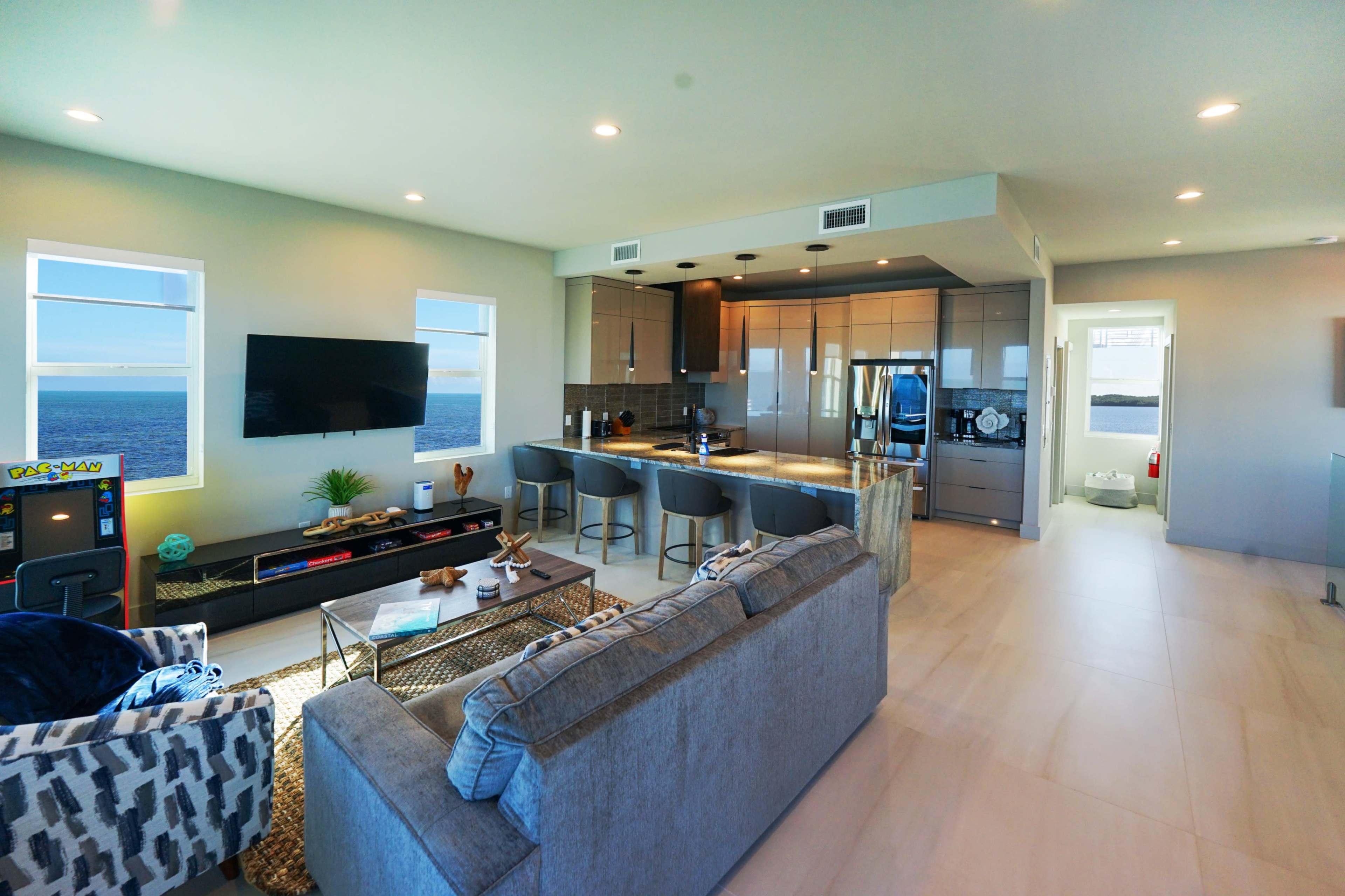 The image shows a modern open-concept living area with a kitchen featuring stainless steel appliances, a dining space with barstools, and large windows overlooking the ocean.