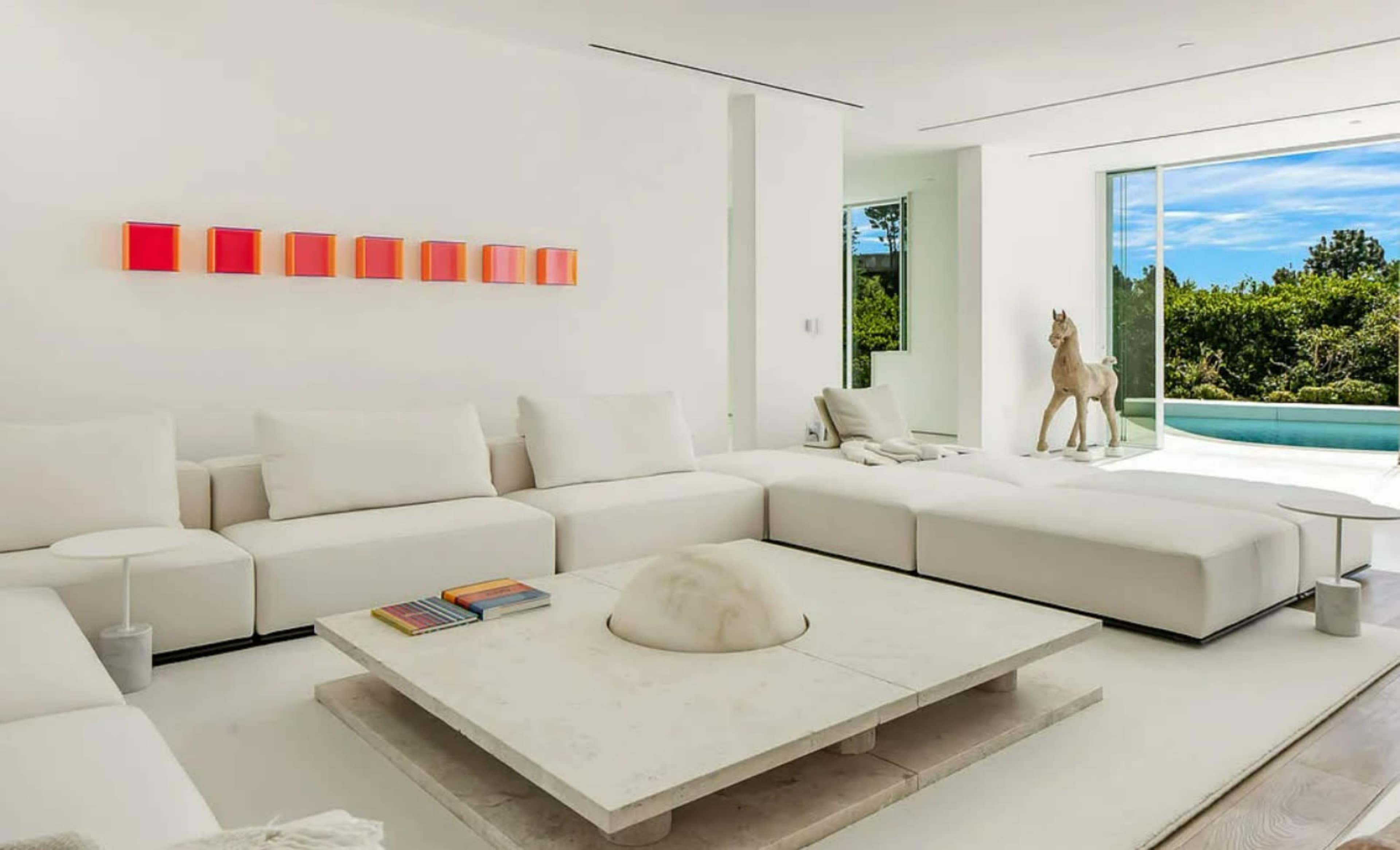 A modern living room features a white sectional sofa, a stone coffee table, and a large window overlooking greenery.