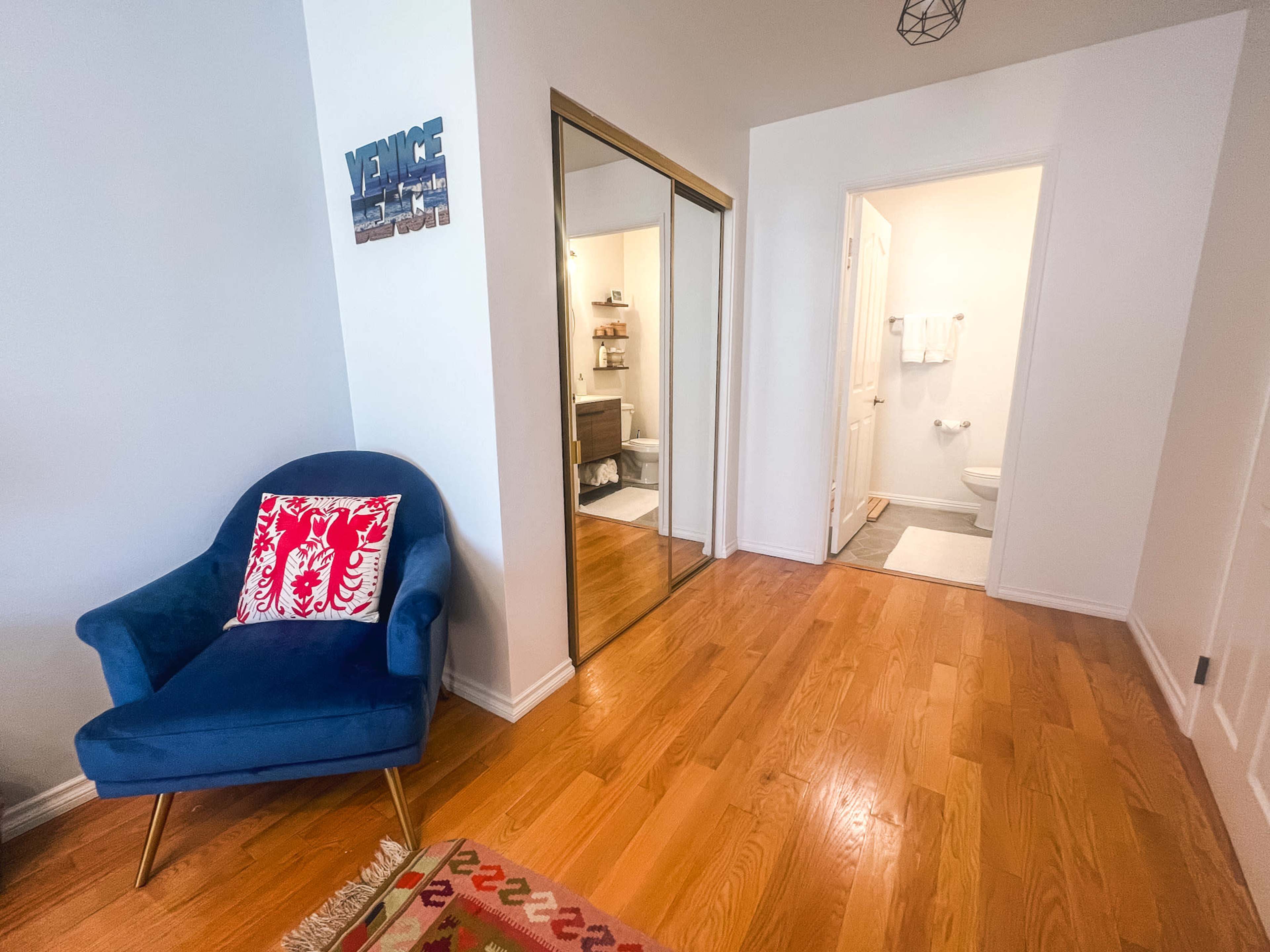 The image shows a small room with hardwood floors, featuring a blue armchair with a decorative pillow and a mirrored closet door leading to a bathroom.