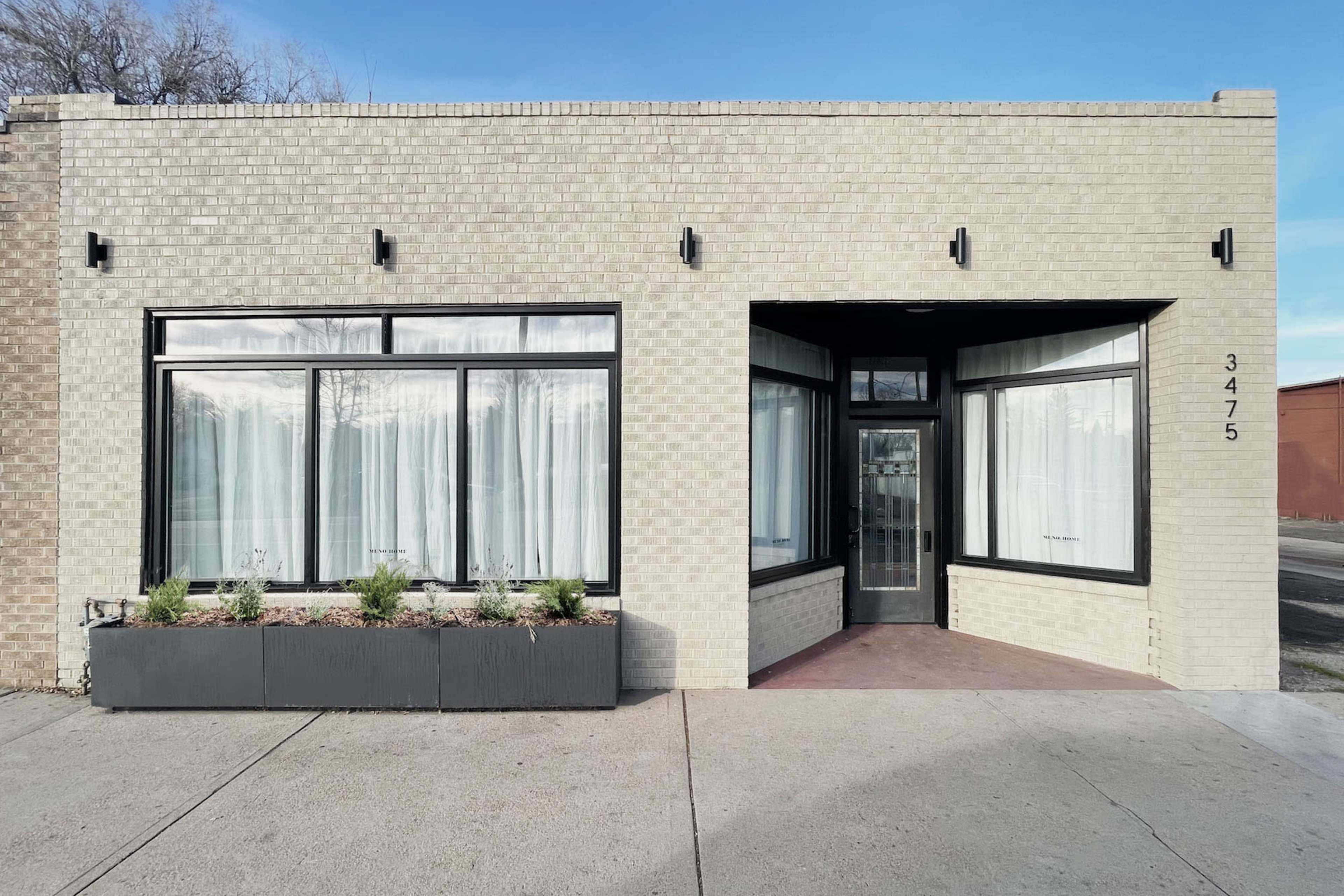 A single-story brick building with large front windows and a centrally located door, flanked by a planter box containing greenery.
