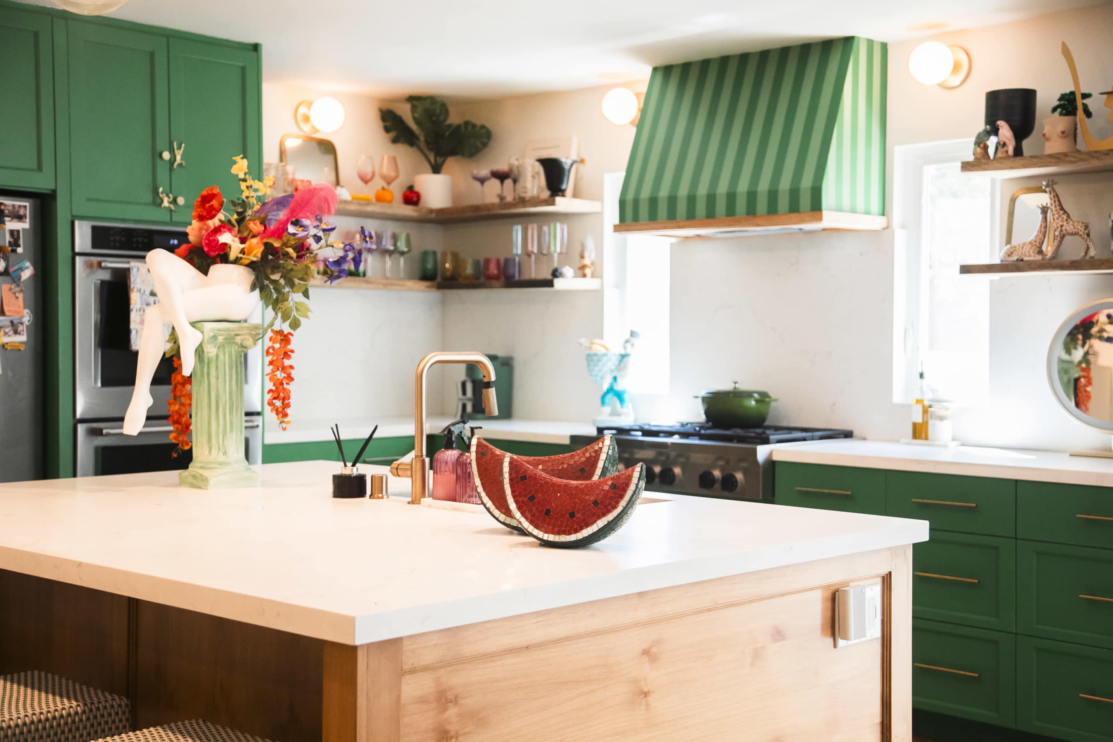 The image shows a vibrant kitchen featuring green cabinetry, a large island with a wooden base, and a variety of decorative items, including a bowl of watermelon on the countertop.