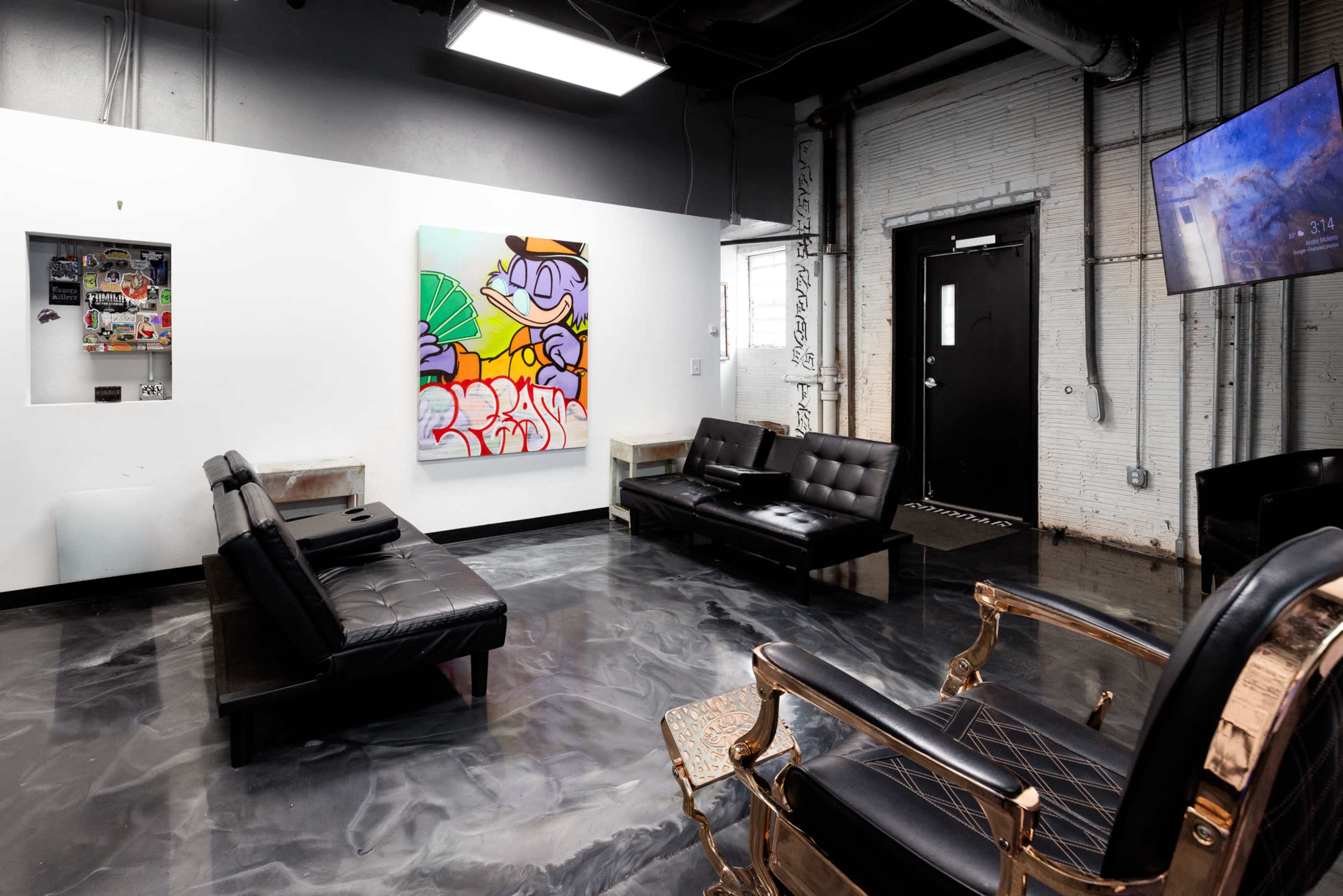 The image shows a modern interior with black leather furniture, a colorful artwork on the wall, and a large screen mounted near a door.