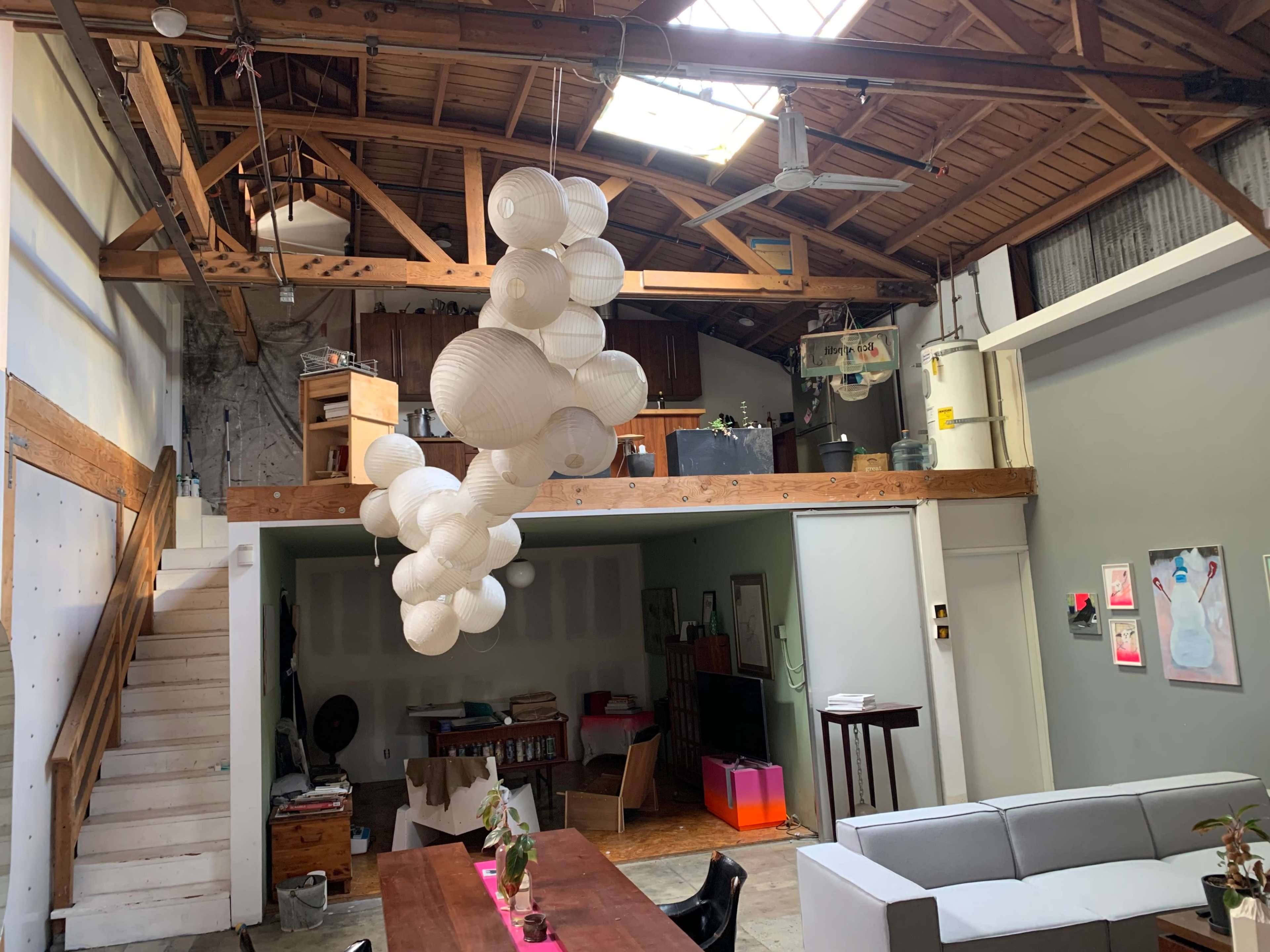 The image shows a spacious loft-style room with high wooden beams, a staircase, and a unique hanging light fixture composed of spherical shapes.