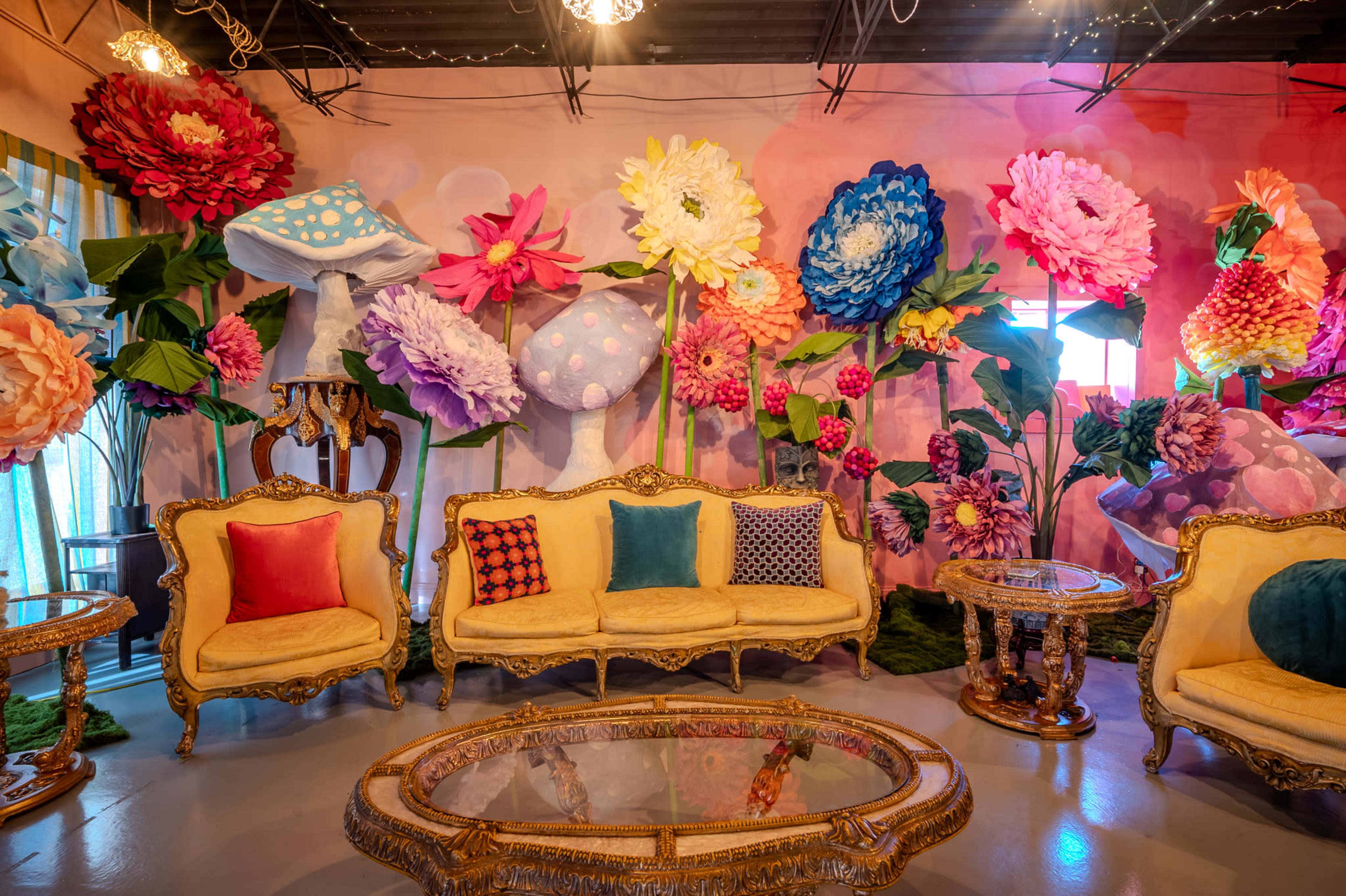 The image shows a vibrant room decorated with large, colorful flower and mushroom motifs on the wall, accompanied by ornate golden furniture and a glass coffee table.