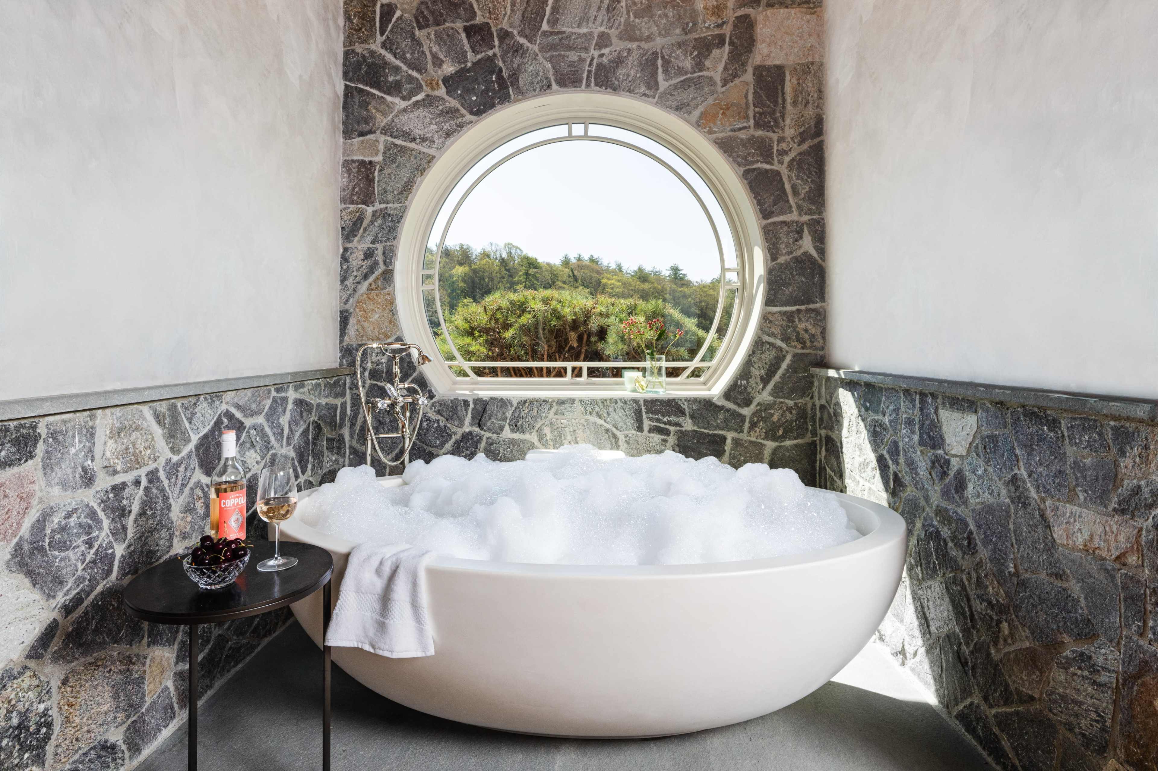 A modern bathroom featuring a freestanding white bathtub filled with bubbles, set against a stone wall with a large round window that overlooks greenery.