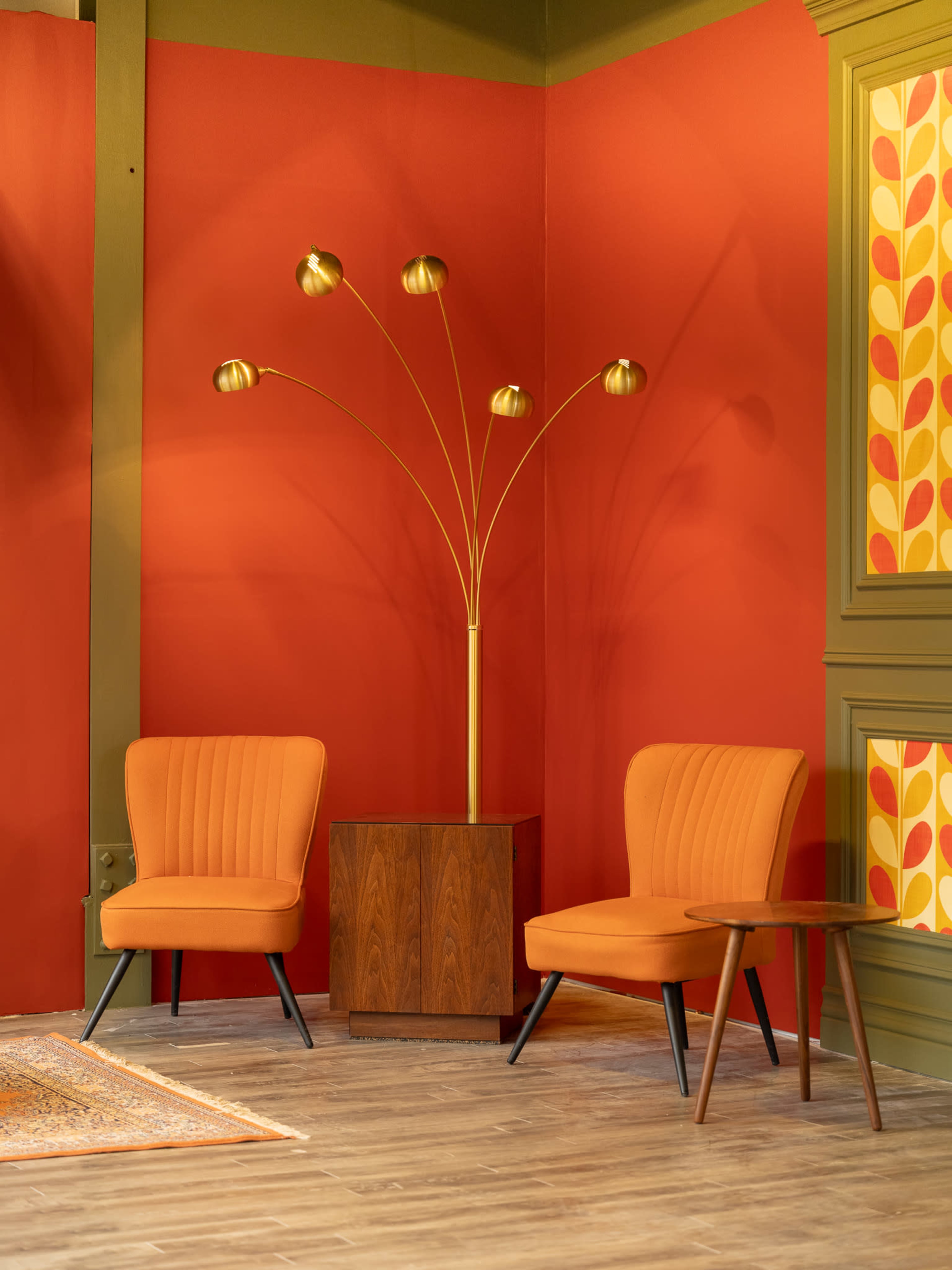 The scene features two orange mid-century modern chairs and a wooden side table beside a tall, gold floor lamp against a vibrant red wall.