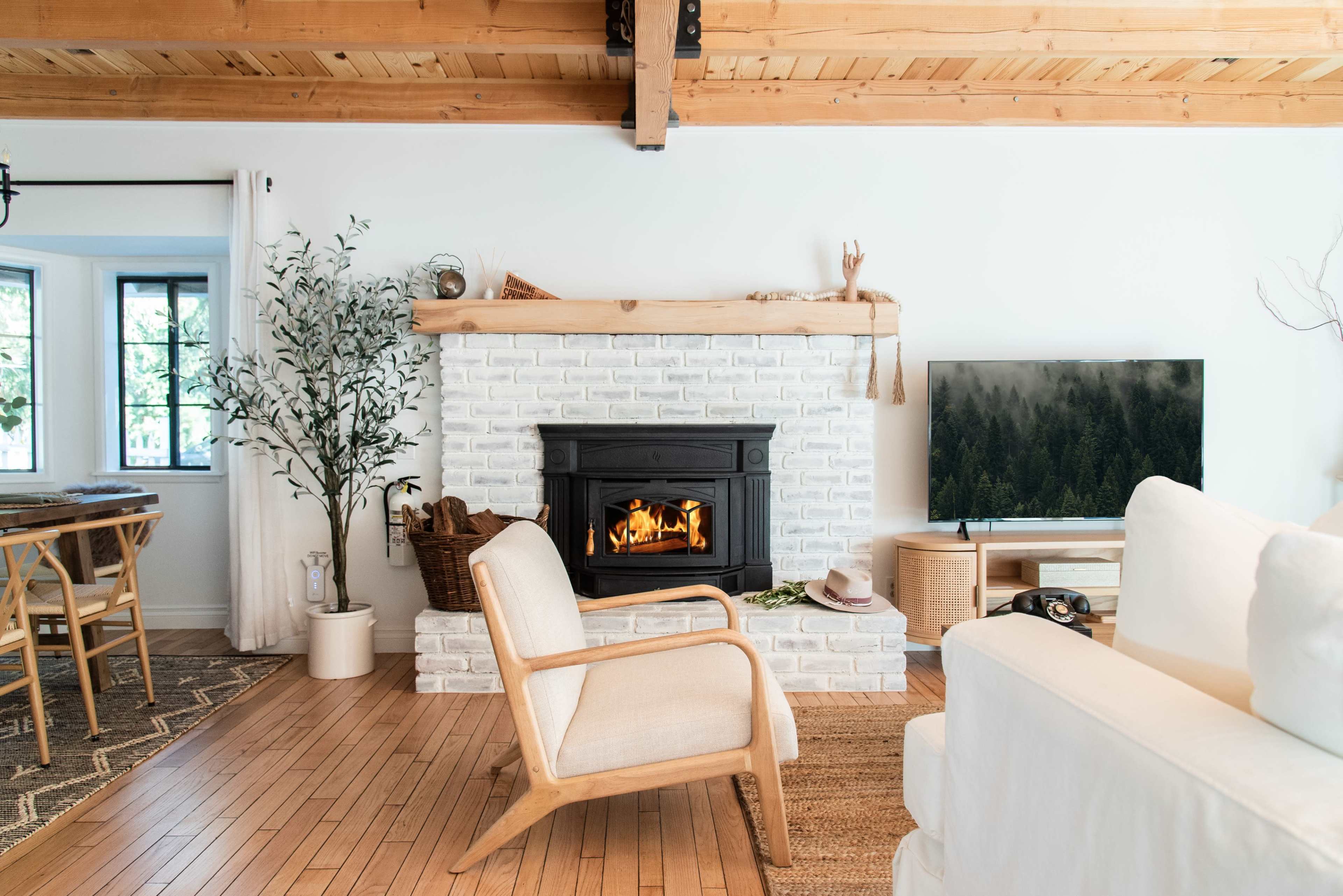 A cozy living room features a white brick fireplace, a television, and wooden accents with a dining area visible in the background.