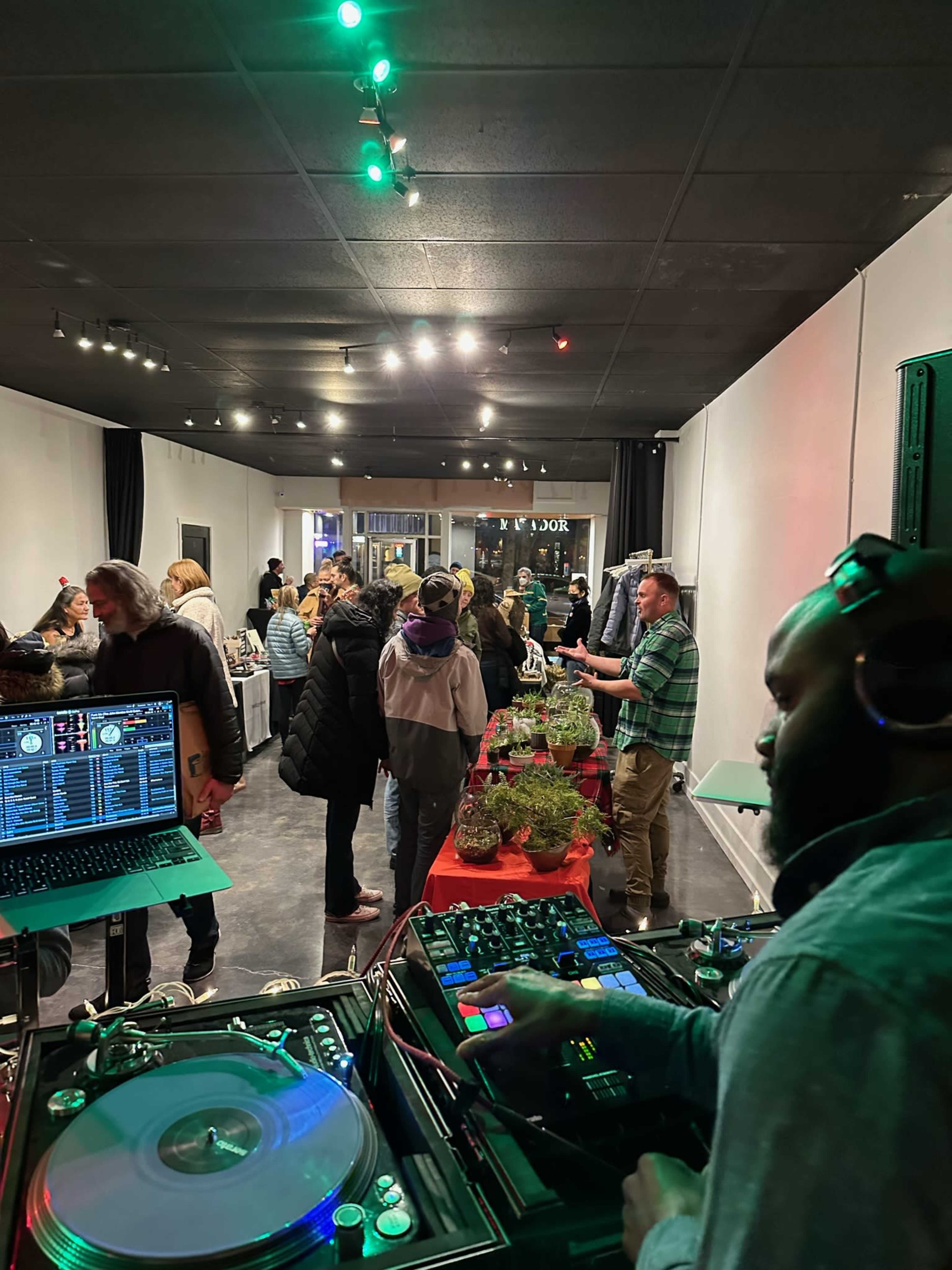 A DJ is mixing music at a crowded indoor event with attendees browsing tables filled with products.