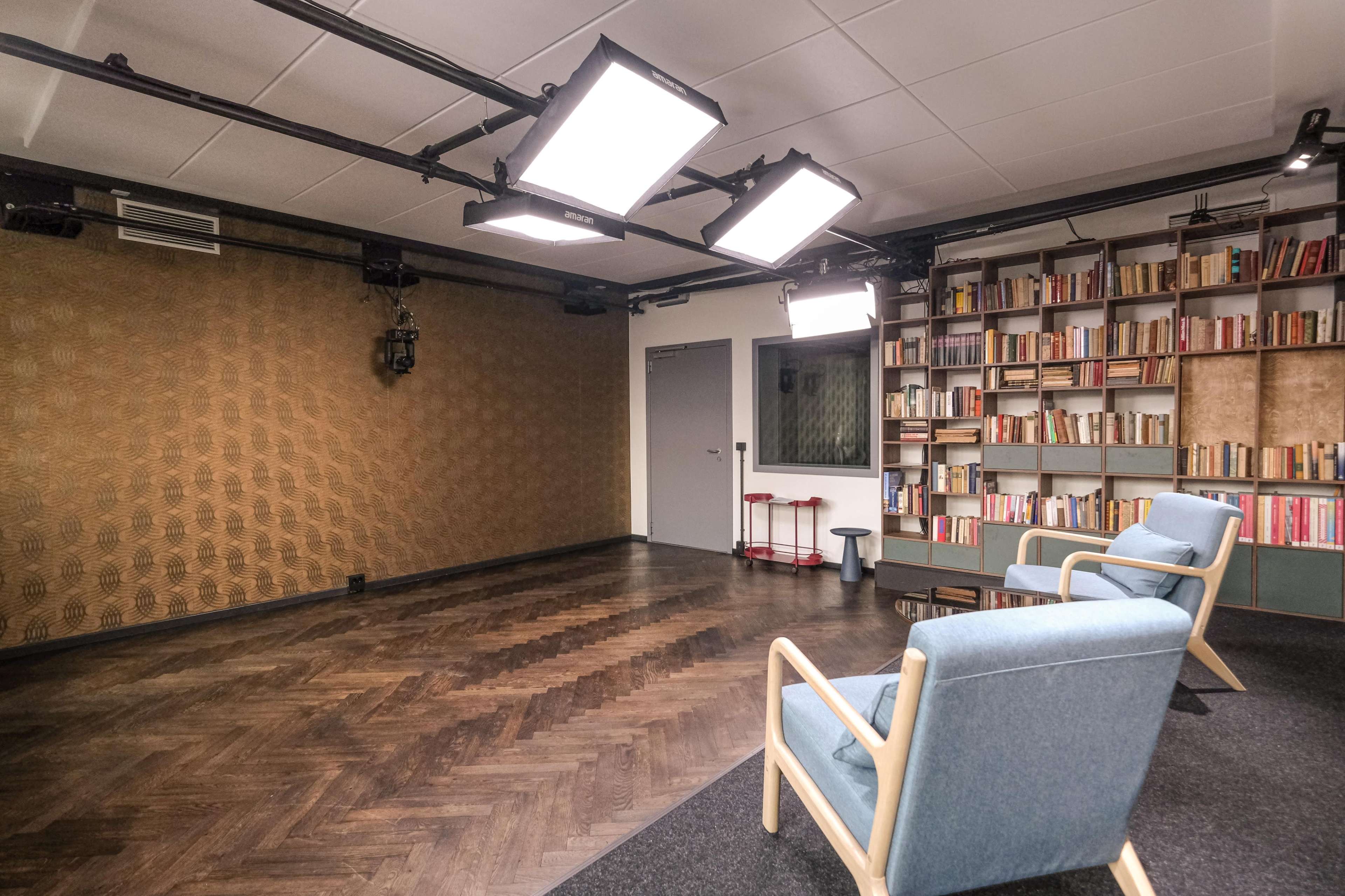 The image depicts a modern studio space featuring two light blue chairs facing a bookshelf filled with books, against a backdrop of textured wall and ceiling lights.