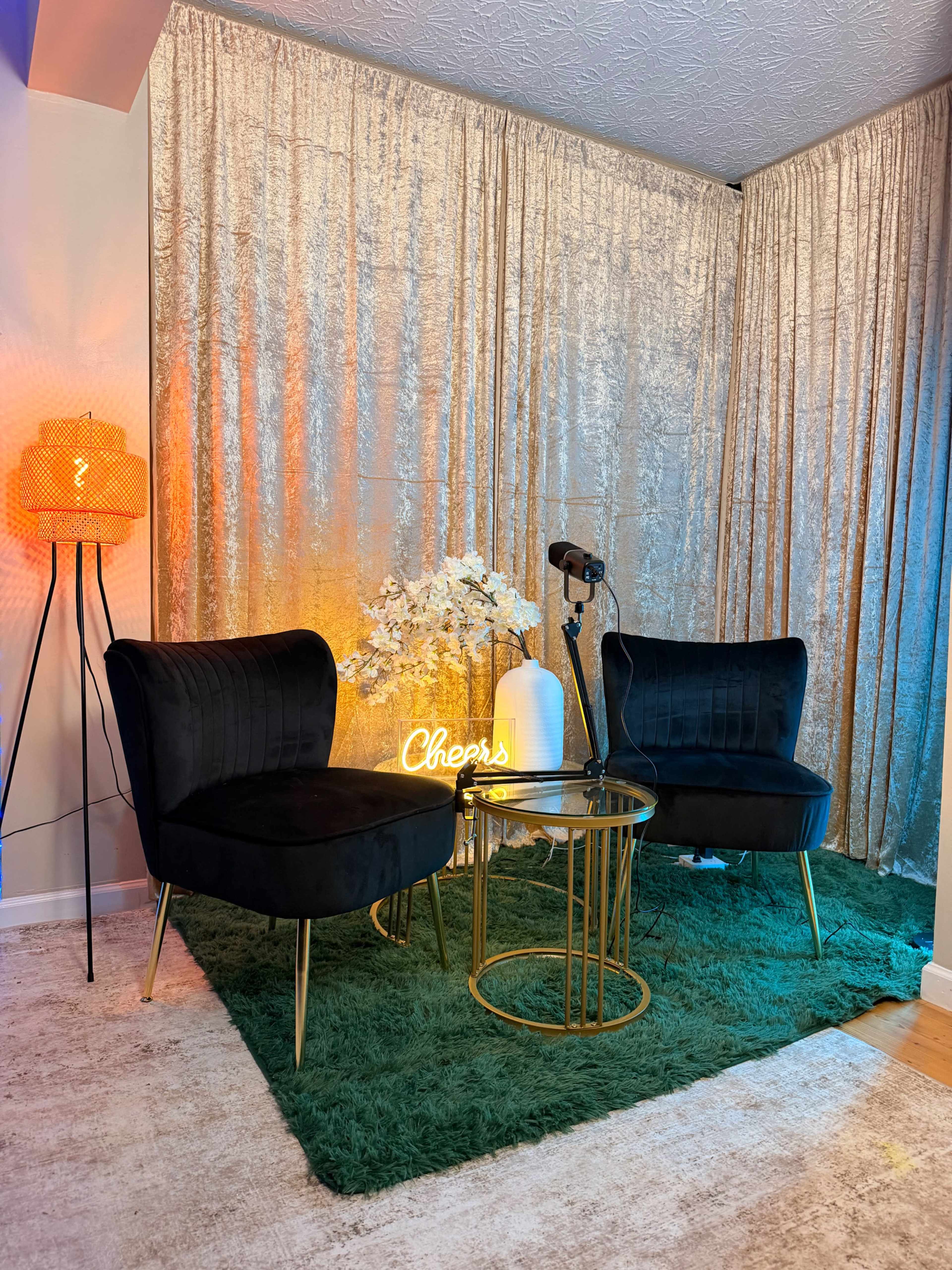 The image features a cozy seating area with two black chairs, a small round gold table, and decorative elements including a neon "Cheers" sign, a flower arrangement, and a floor lamp, all set against a backdrop of shimmering metallic curtains.