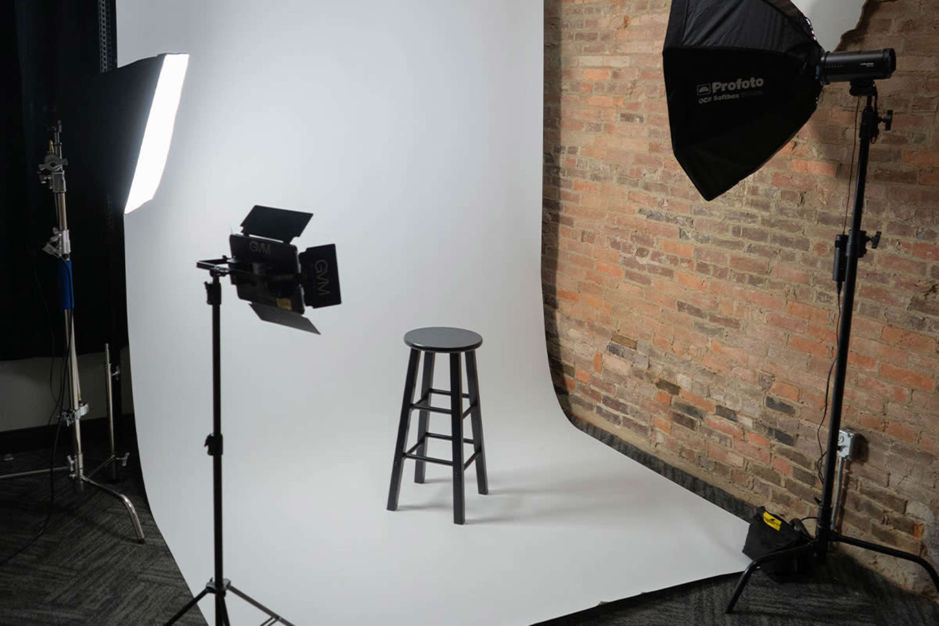 A black stool is positioned on a white backdrop with soft lighting equipment on either side.