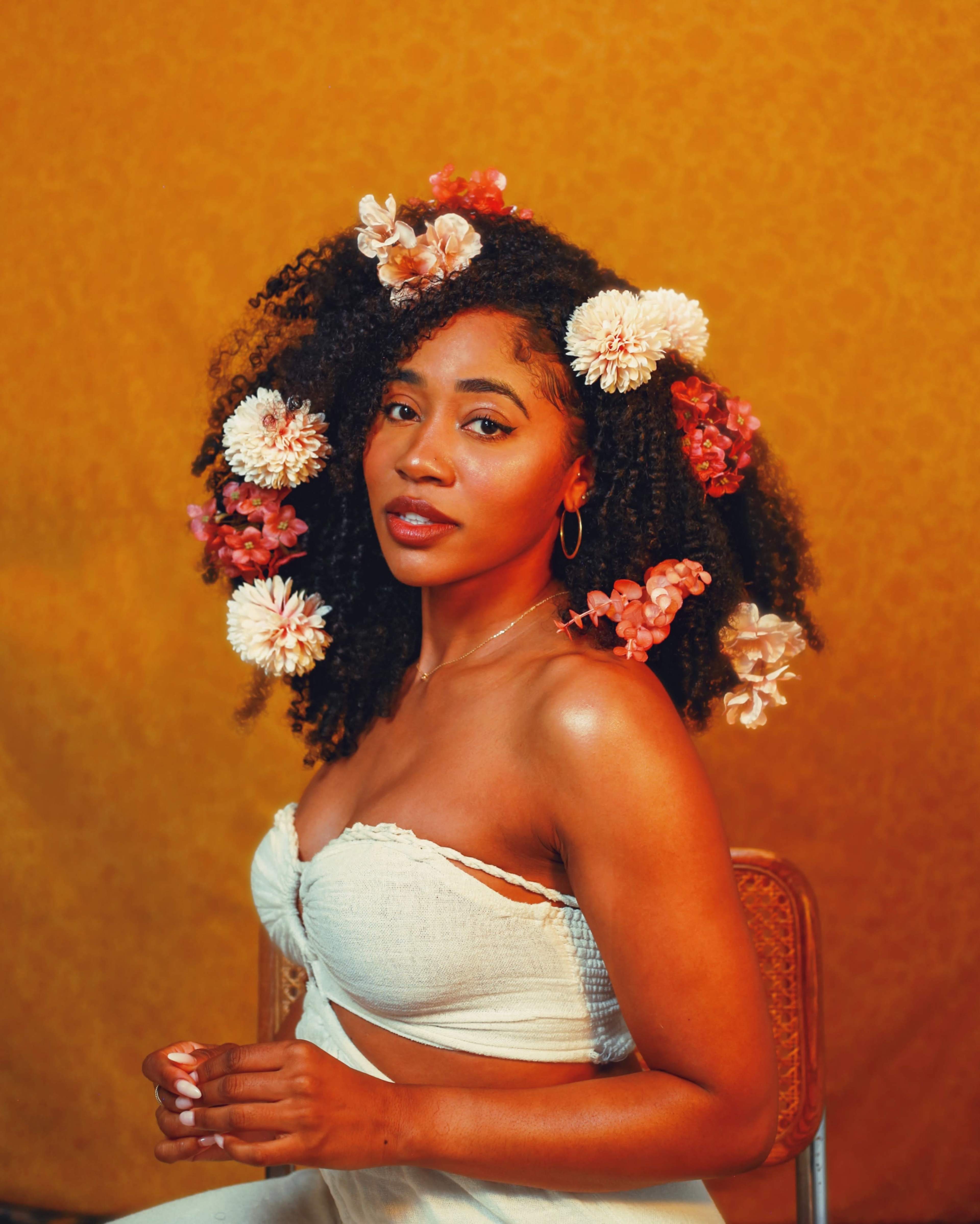 A woman with curly hair adorned with flowers poses confidently against a textured yellow background.