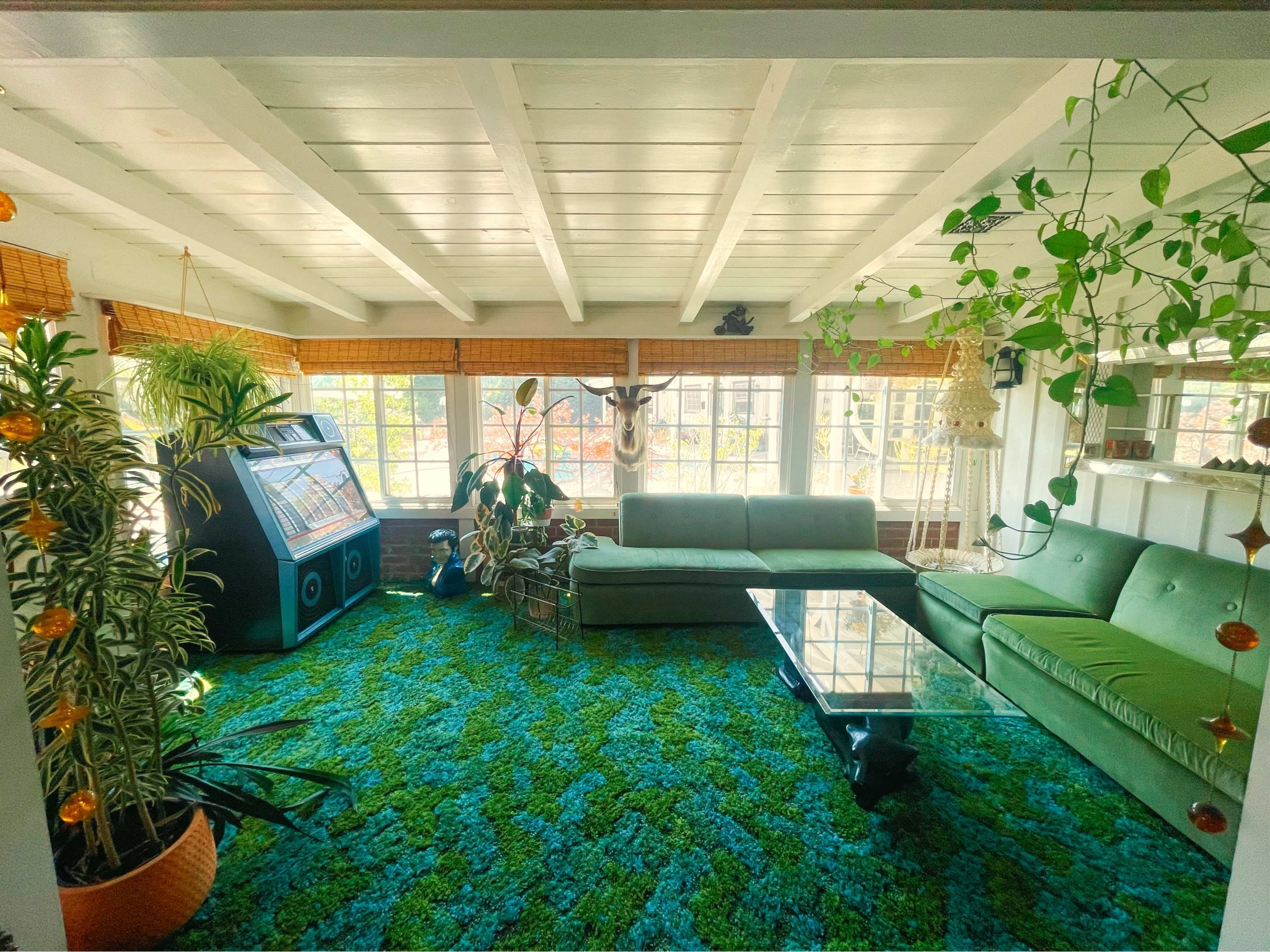 The room features a green carpet, a glass coffee table, a large sofa, and a vintage jukebox, all illuminated by natural light from the windows.