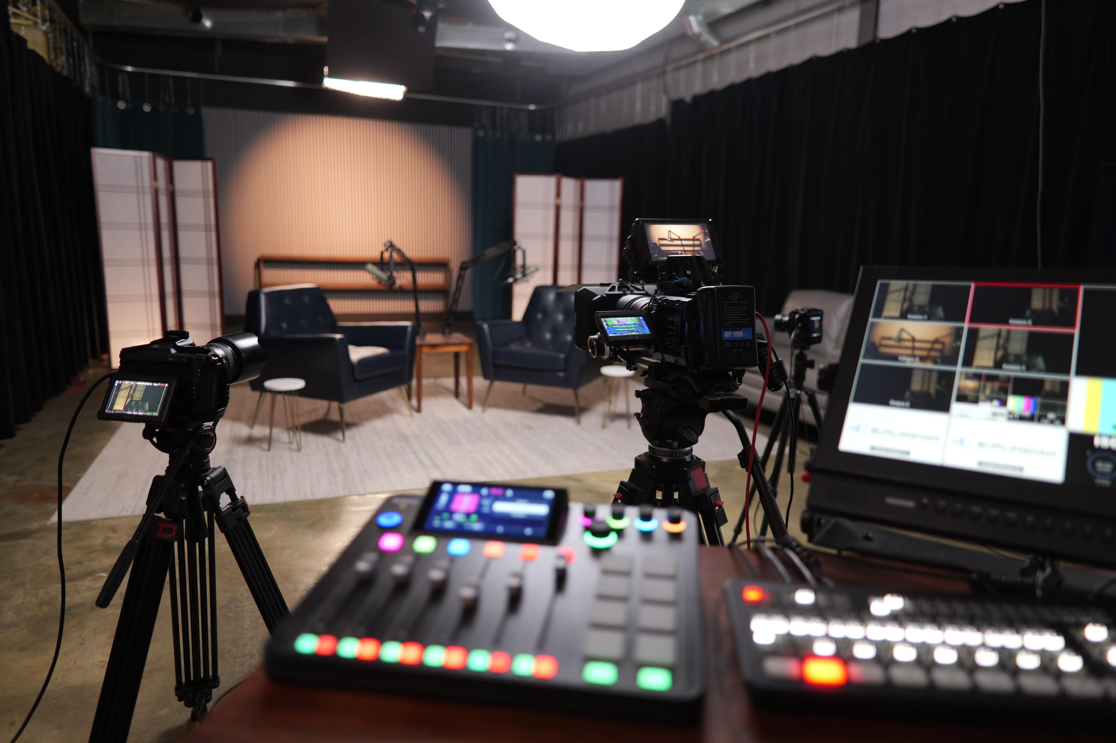 The image shows a multimedia studio setup with cameras, a control panel, and a seating arrangement in the background.