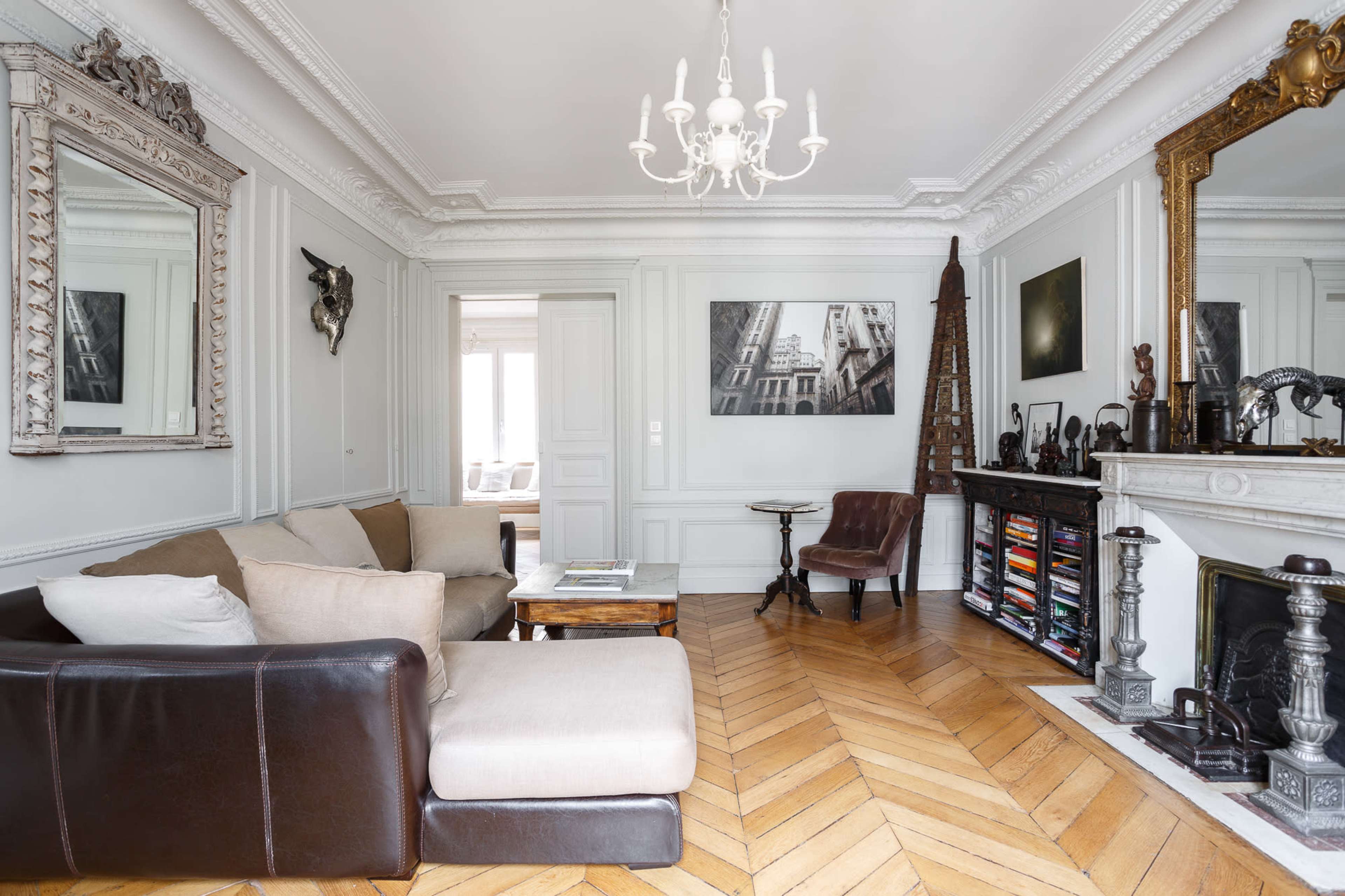 The living room features a sectional sofa, a vintage armchair, a large mirror, and a bookshelf filled with books, all set against intricately paneled walls and hardwood flooring.
