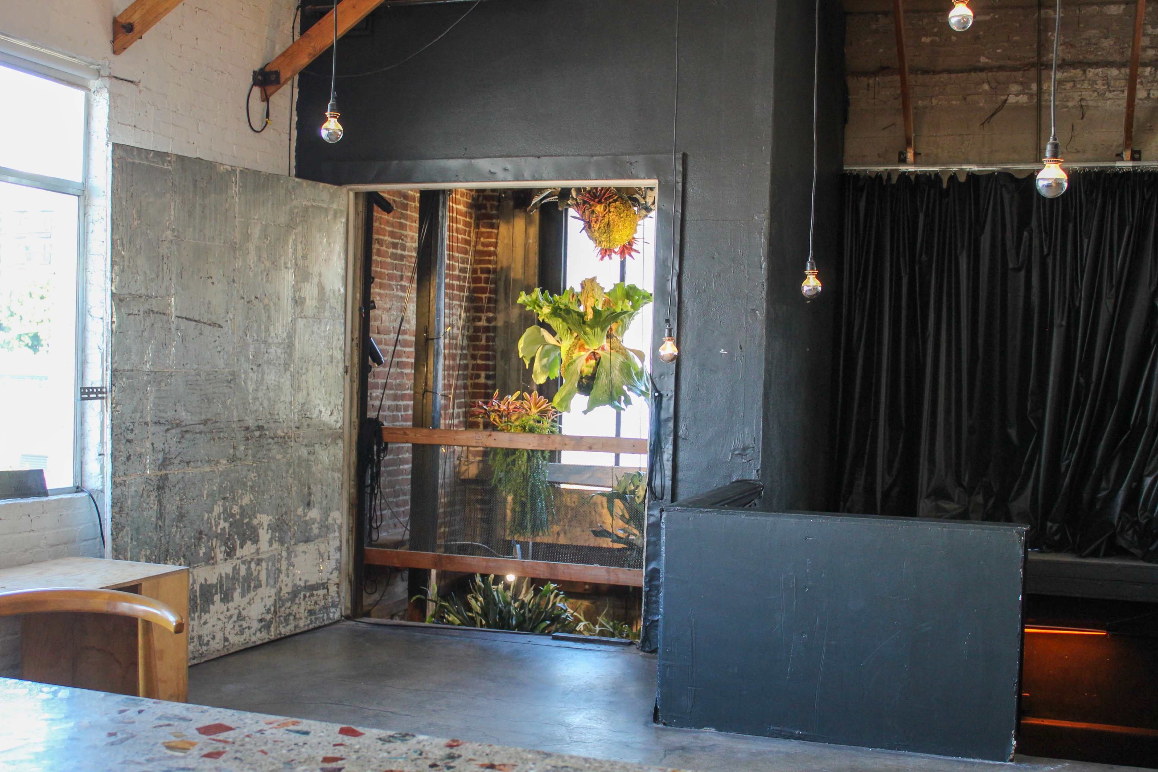 The image shows a minimalist interior space with concrete walls, wooden beams, and a large opening that leads to a greenery-filled area.