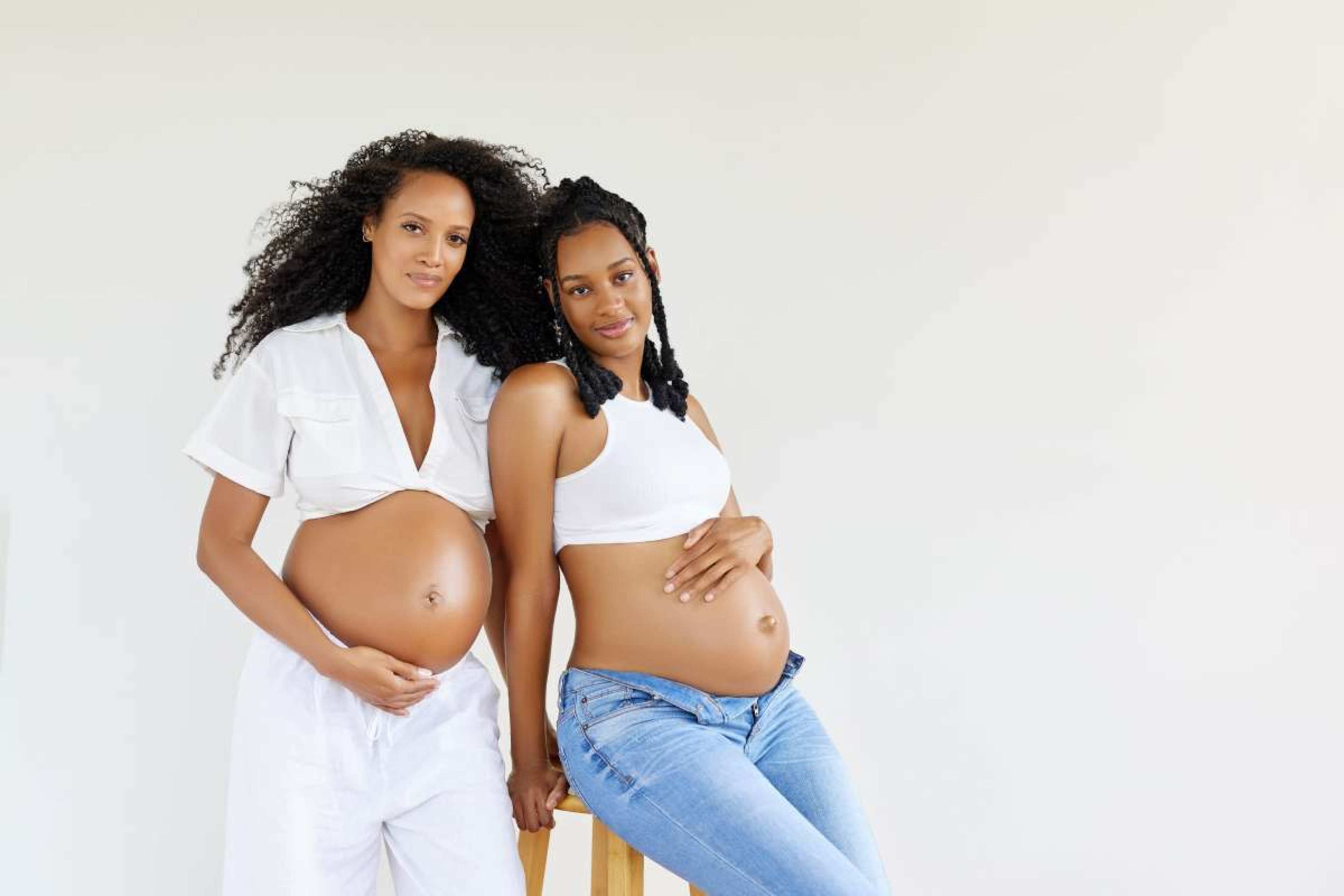 Two pregnant women pose side by side, one wearing a white outfit and the other in a white top with jeans, both showing their baby bumps.