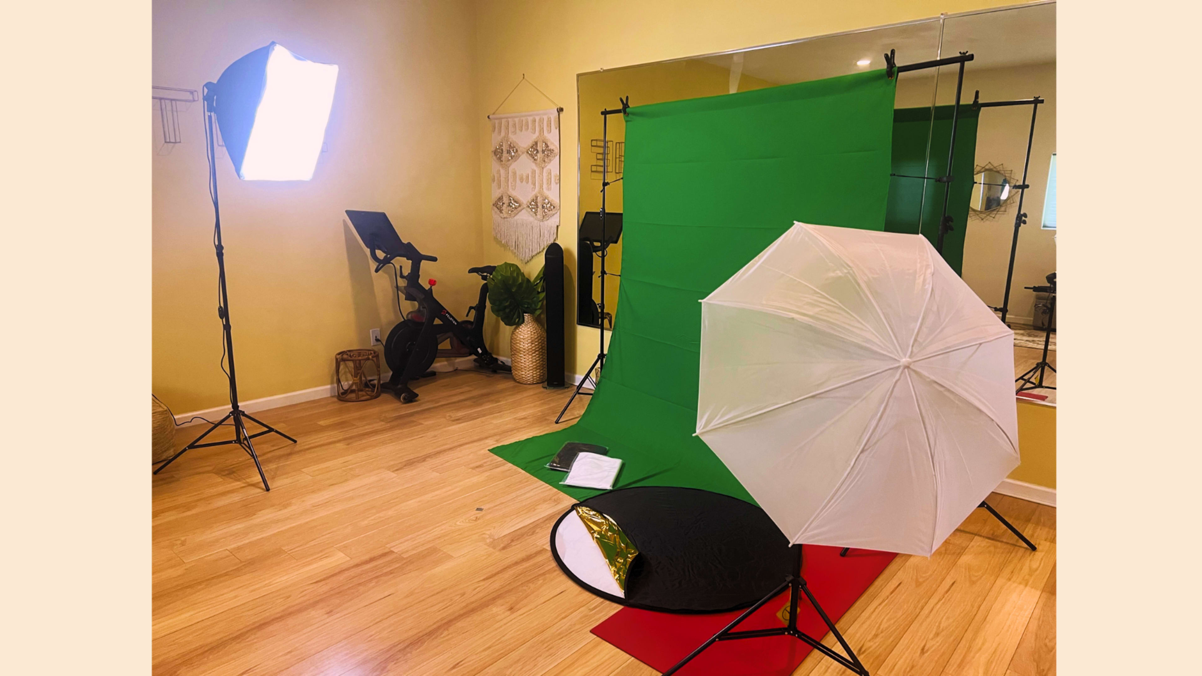 A photography setup with a green backdrop, lighting equipment, an umbrella, and a stationary bike in a brightly lit room.