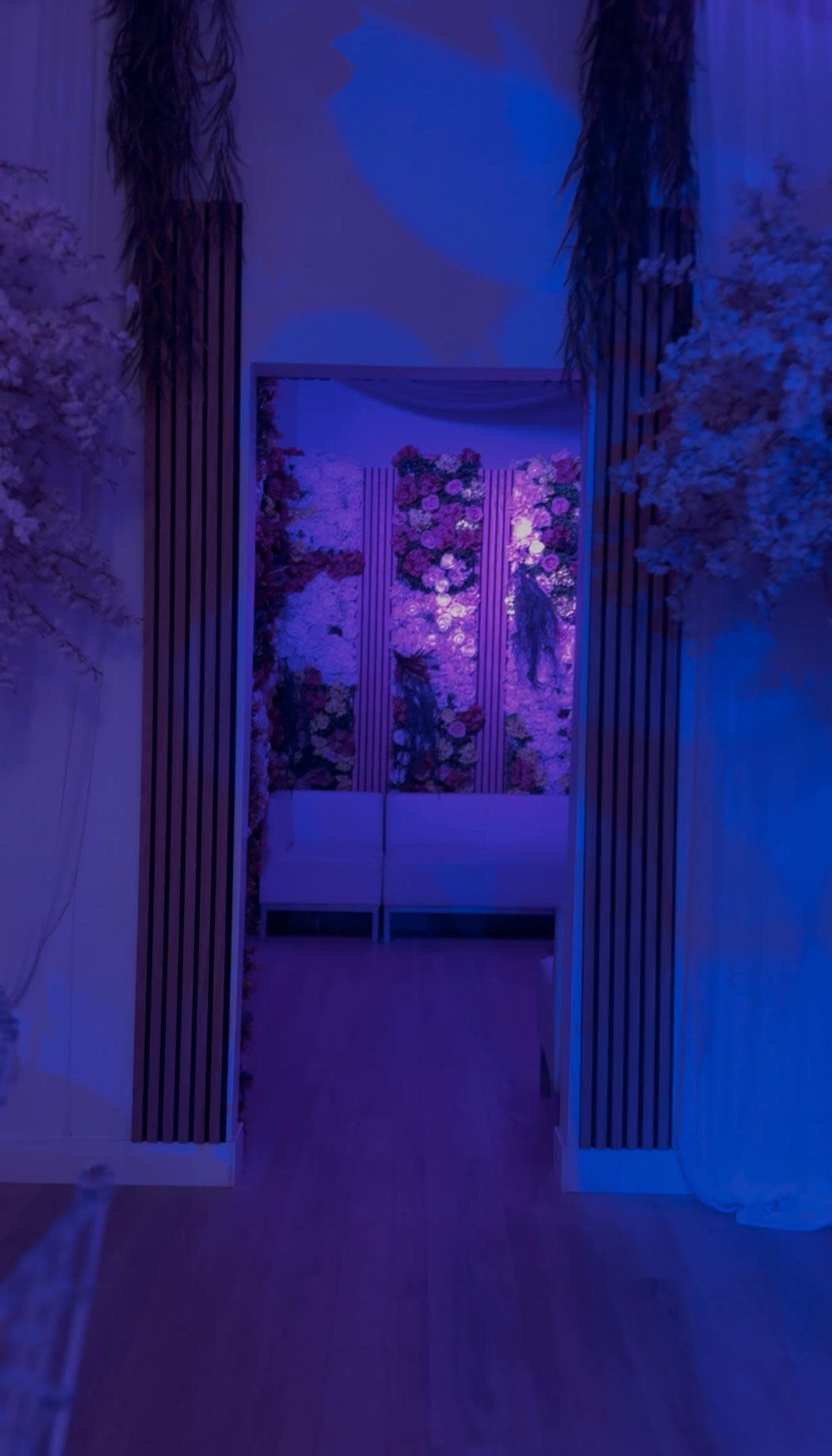 A hallway leads to a softly lit area adorned with floral decorations and illuminated in shades of purple.