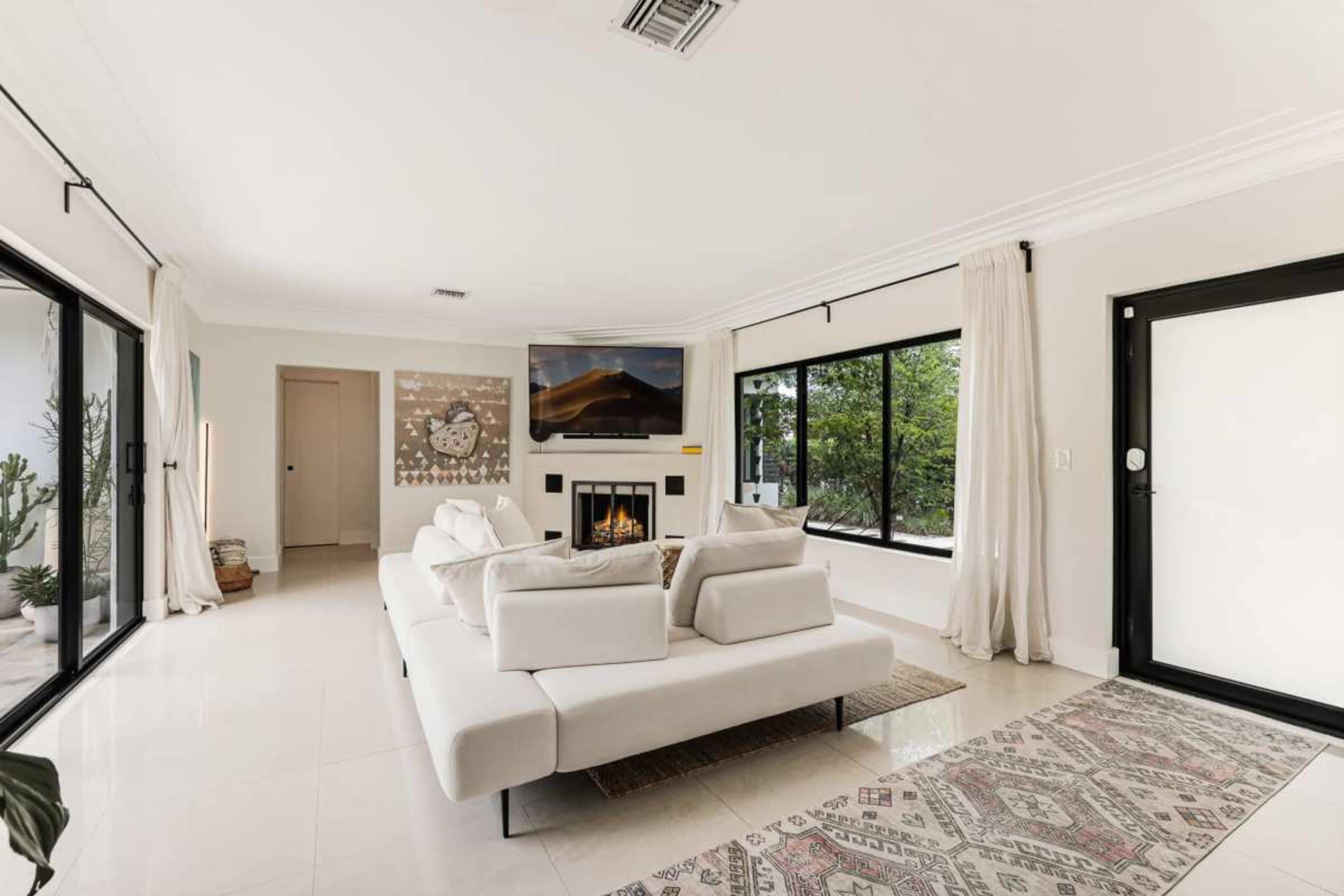 A modern living room features a light-colored sectional sofa, a flat-screen TV above a fireplace, large windows with natural light, and minimalistic decor.