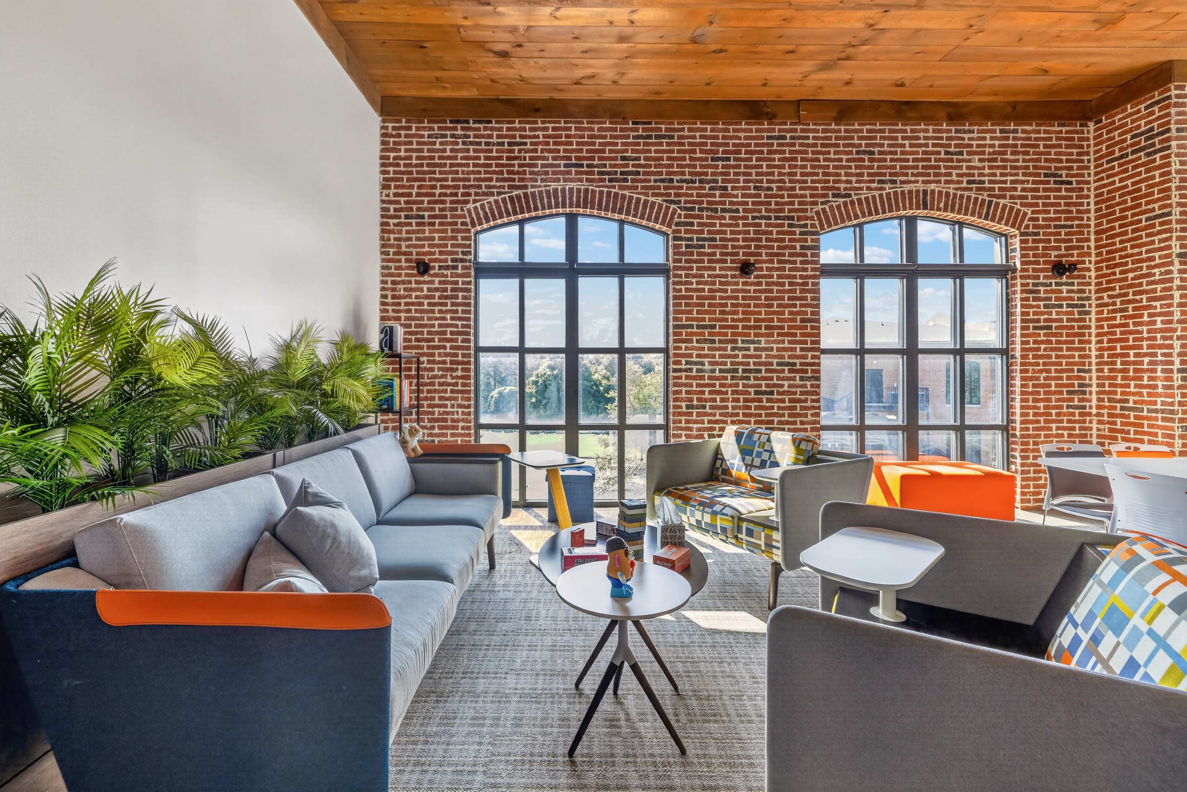The image features a modern lounge area with large arched windows, brick walls, and contemporary seating arrangements centered around a coffee table.