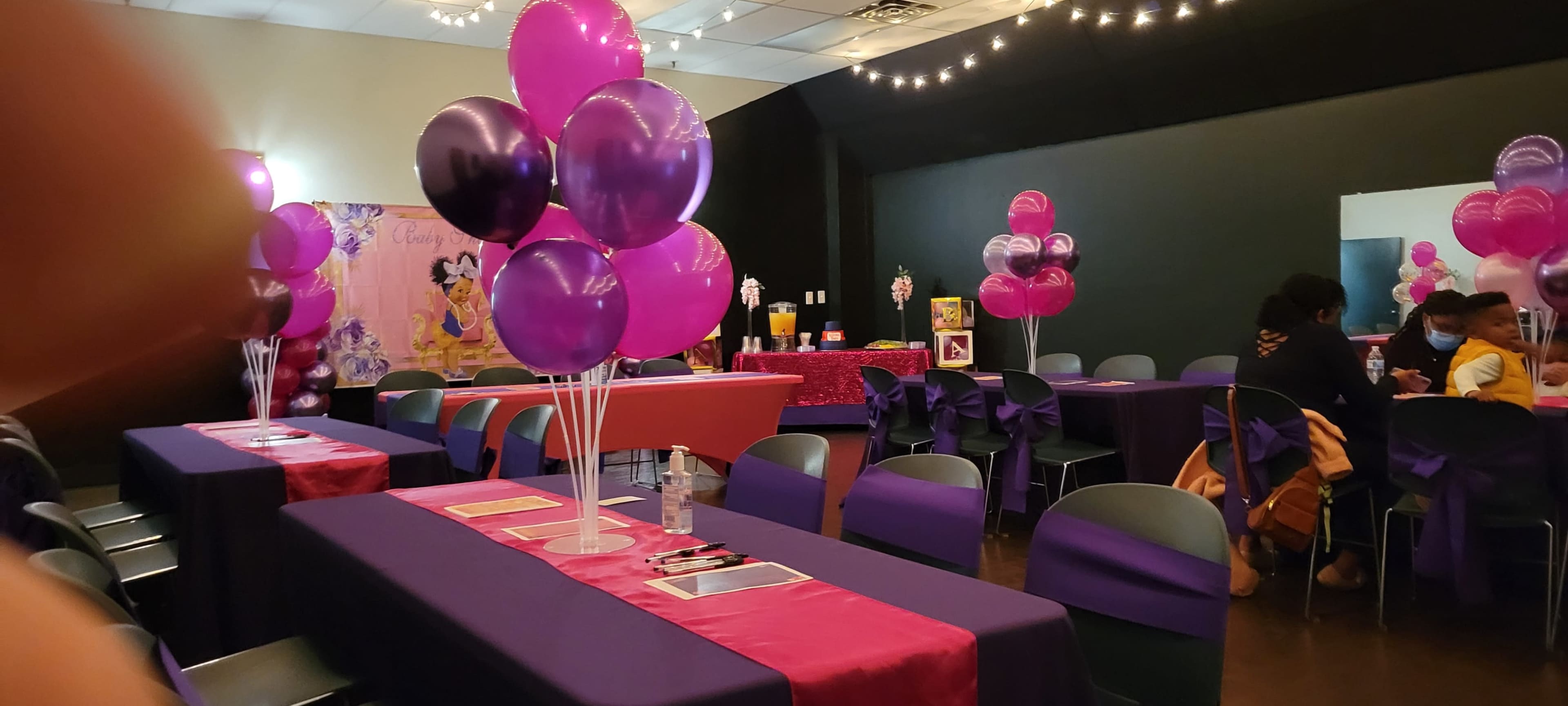 The image shows a decorated event space with purple and pink balloons, tables covered in matching cloths, and a backdrop featuring a character theme for a celebration.