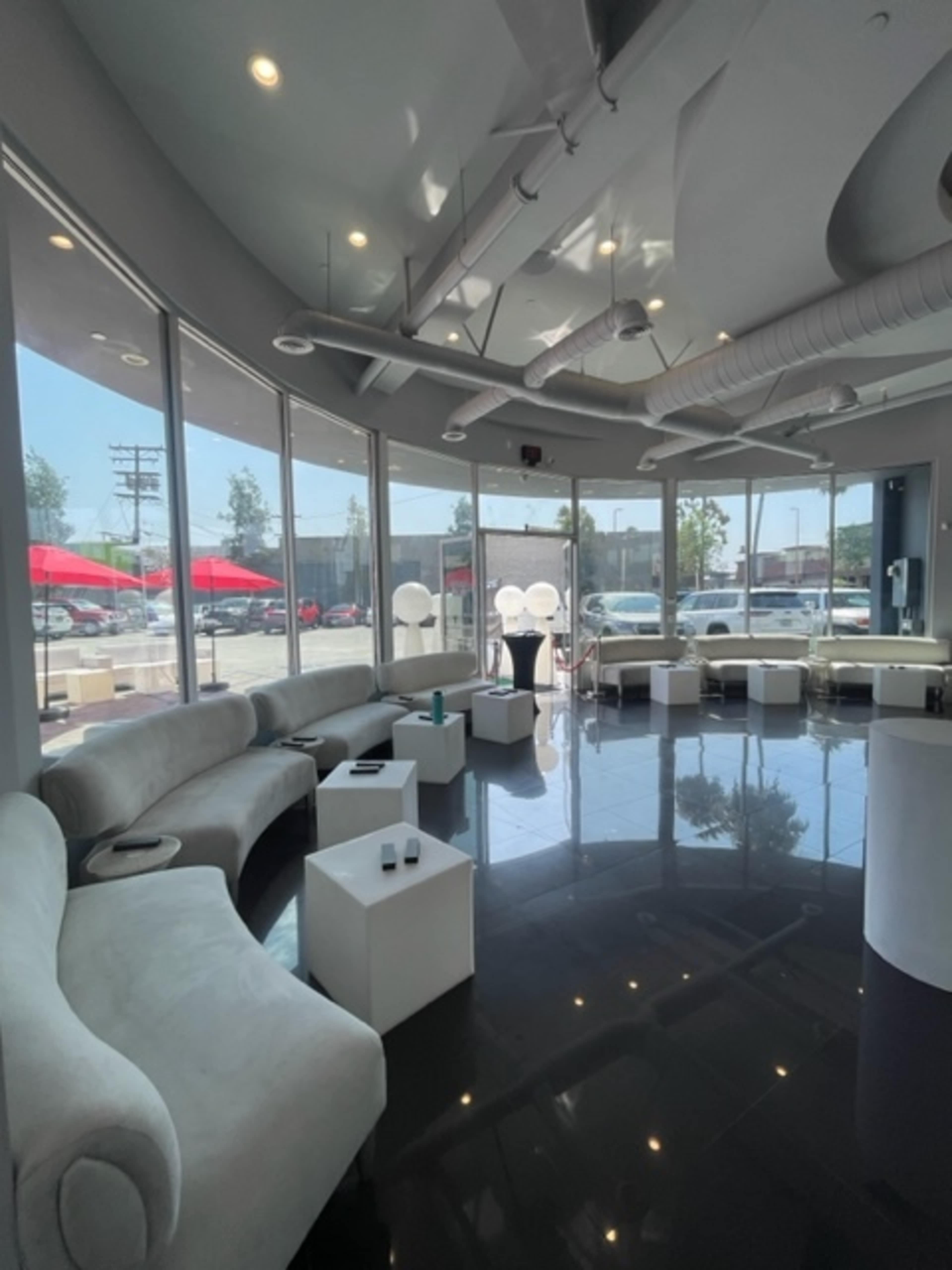 The image shows a modern lounge area with curved white seating, glass walls, and several white tables, illuminated by natural light from large windows.