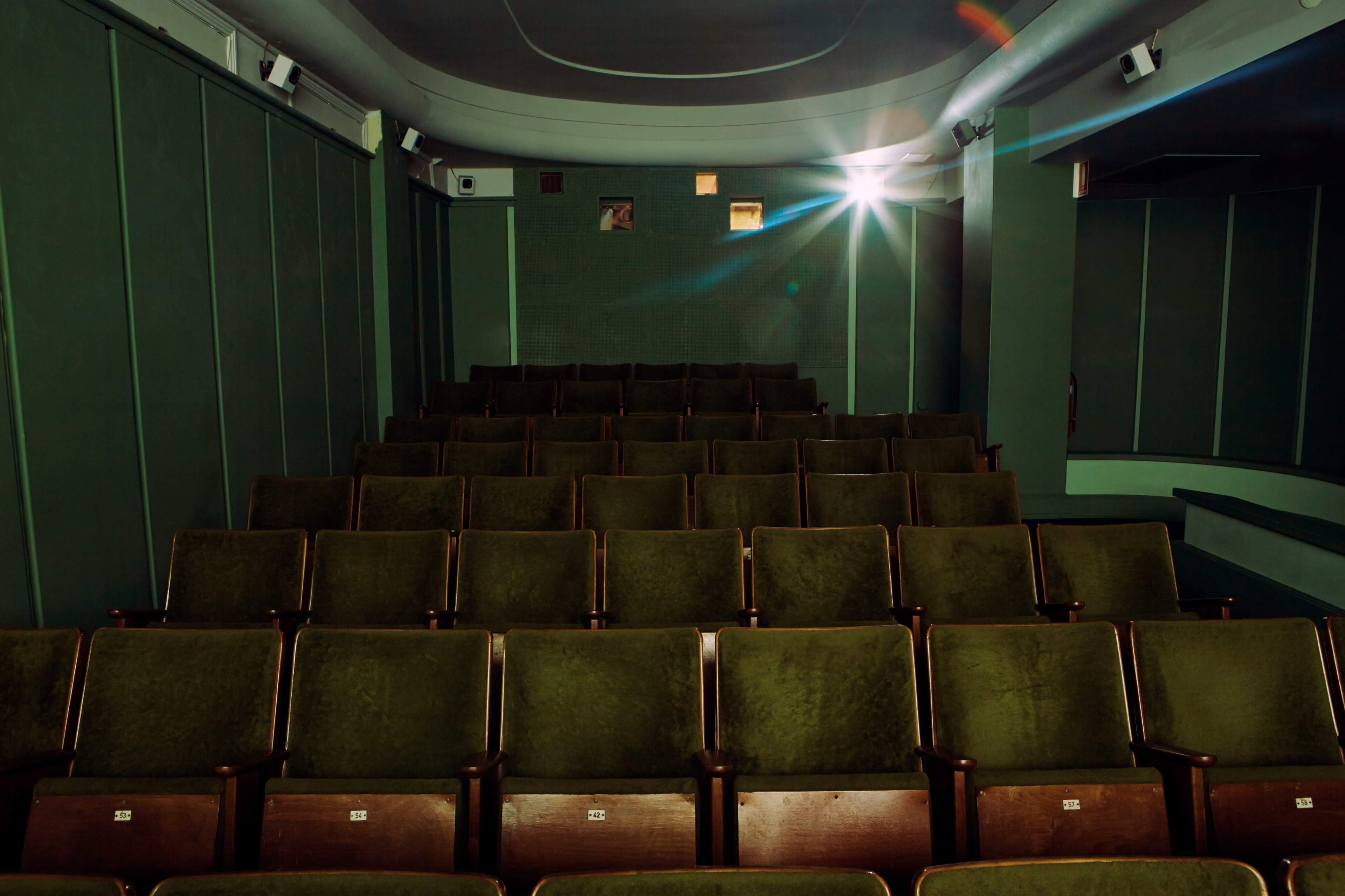 The image shows an empty theater with green upholstered seats arranged in rows, illuminated by a beam of light from the stage.
