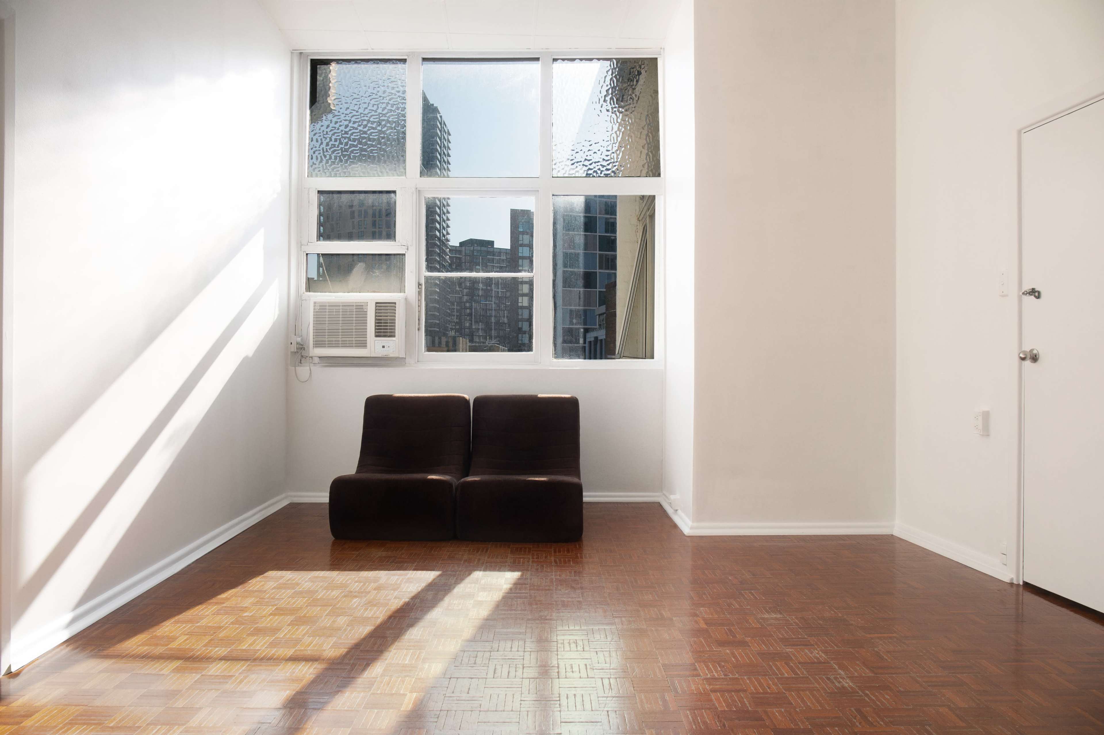 A minimalistic room with a pair of brown chairs, a large window letting in sunlight, and a visible air conditioning unit on the wall.