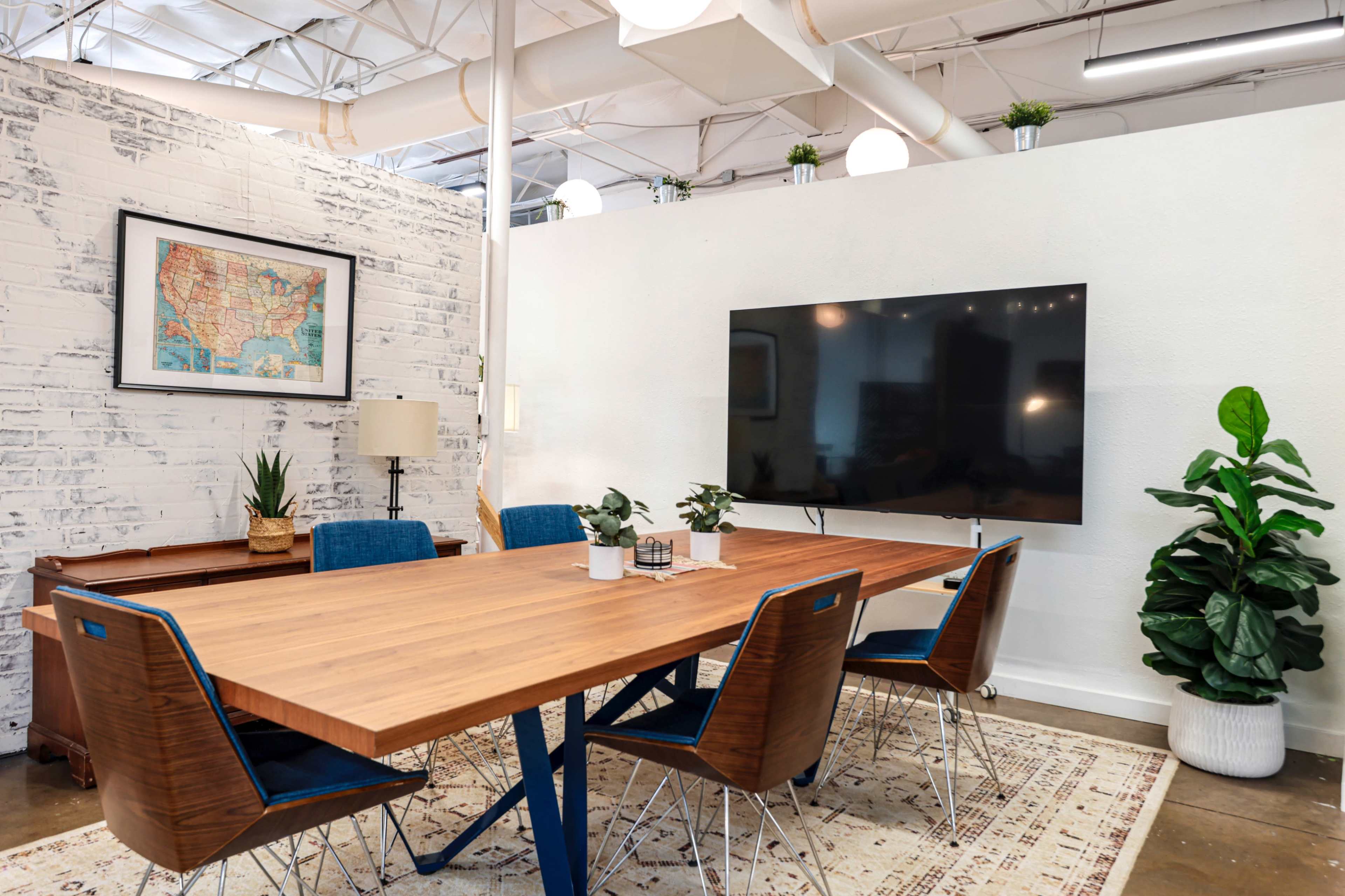 A modern conference room features a wooden table surrounded by six chairs, a wall-mounted television, and a map artwork on the white brick wall.