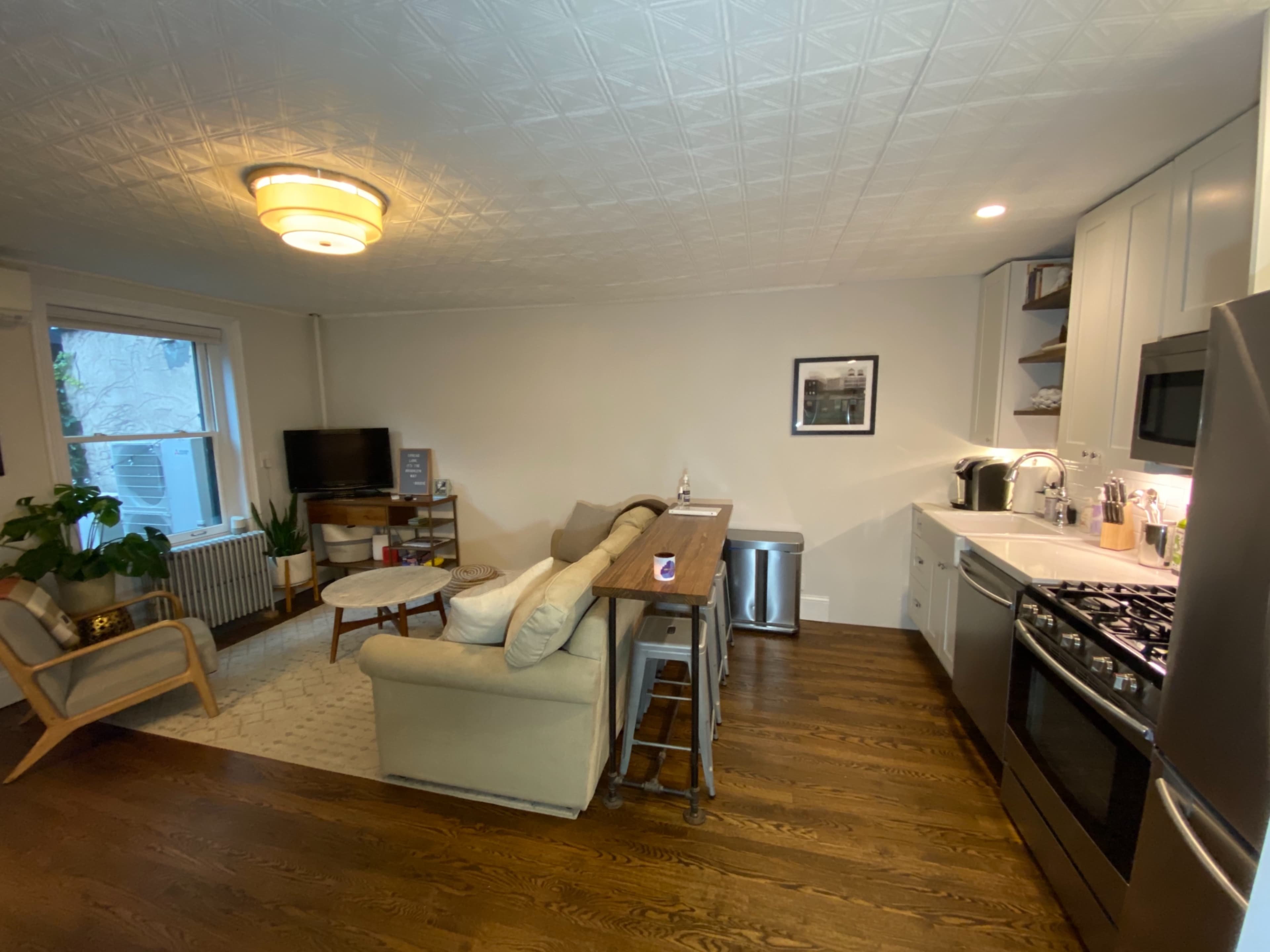 The image shows a modern kitchen and living area with a sofa, a small dining table, a television, and a kitchen counter with appliances.