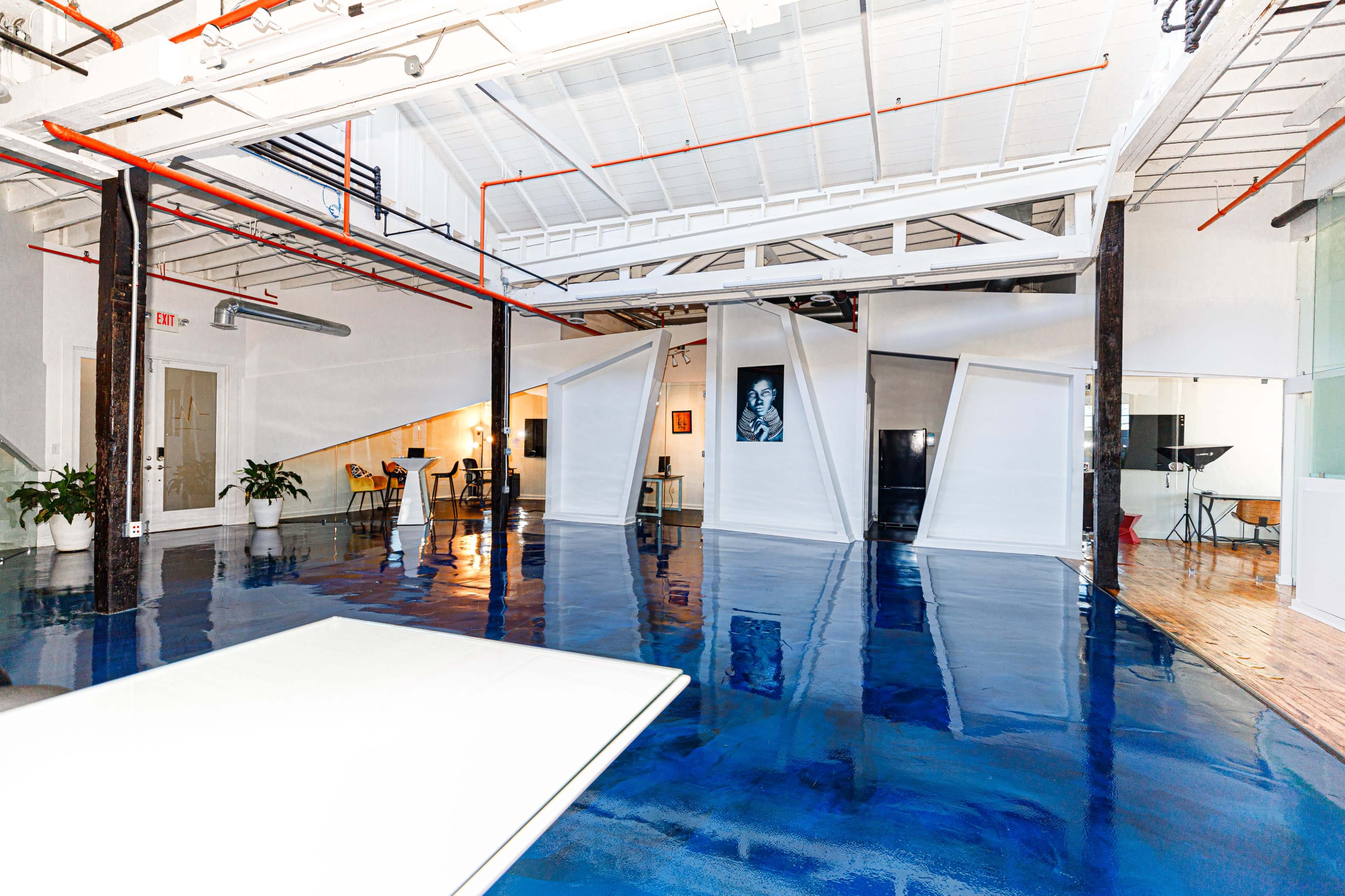 A spacious, modern interior with polished blue floors, white walls, and unique angular partitions, featuring a mix of furniture and greenery.
