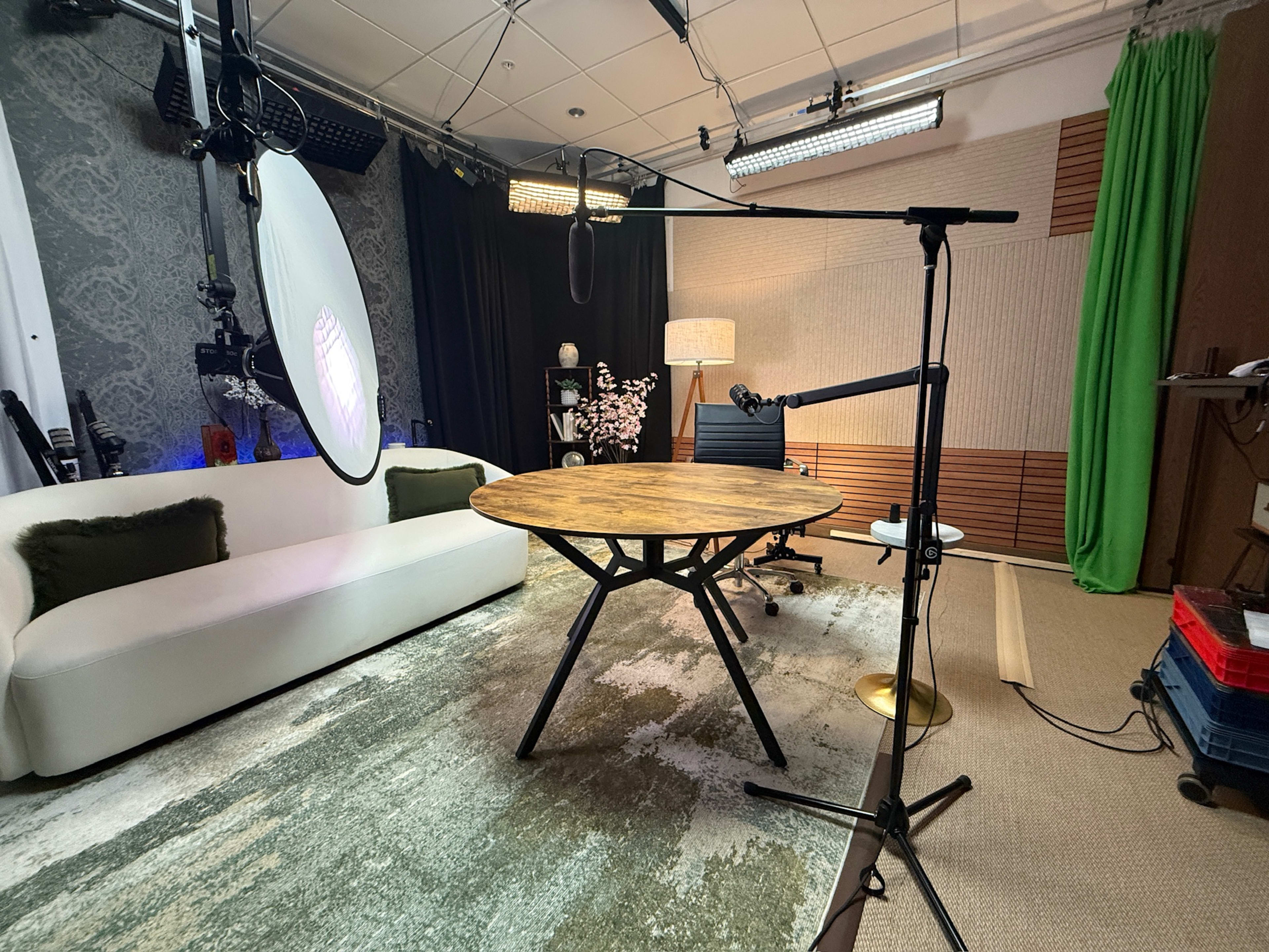 The image shows a media production studio featuring a circular wooden table, a white couch, a microphone setup, and lighting equipment against a backdrop with a green curtain and decorative elements.