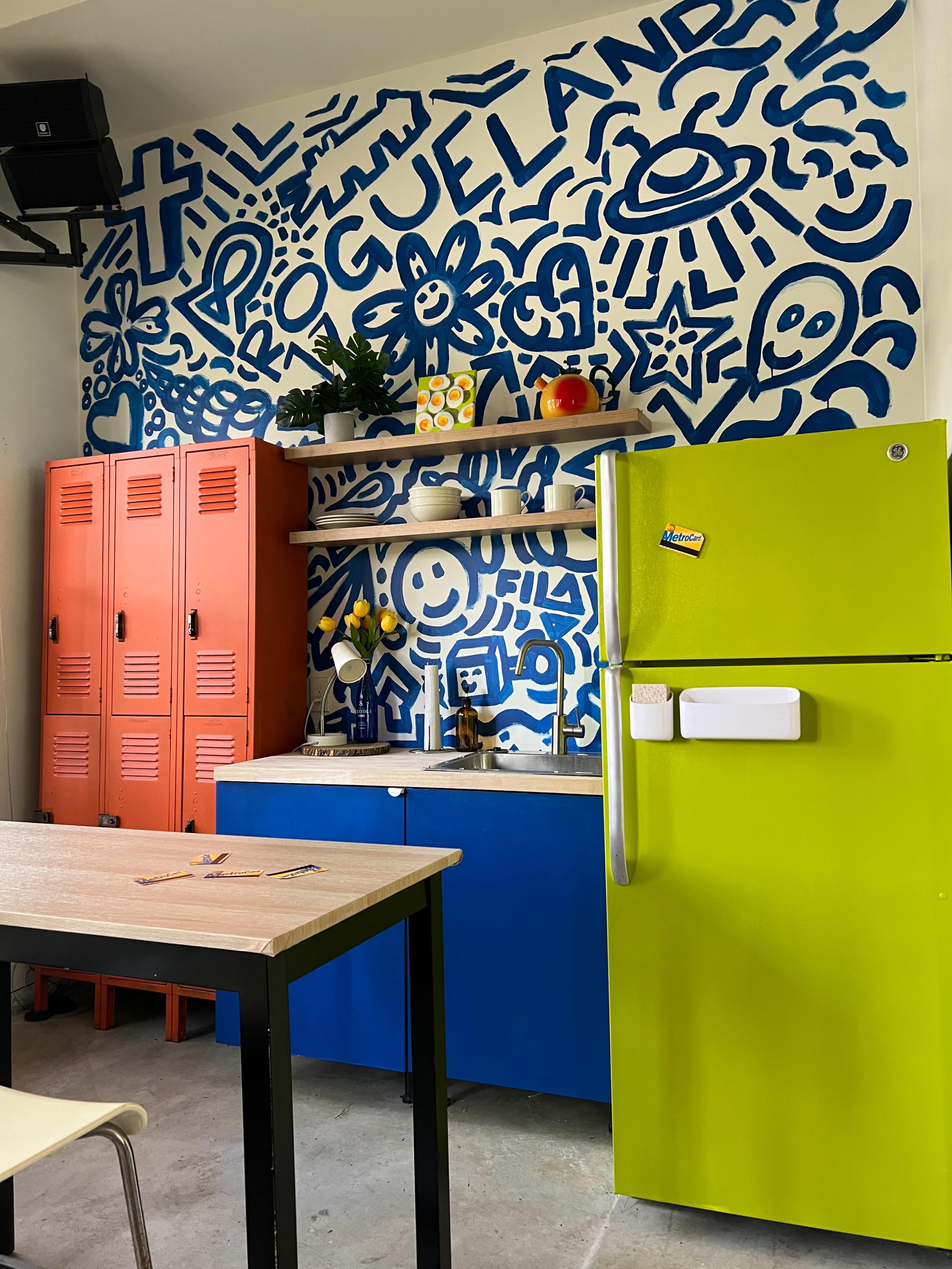 The image shows a vibrant kitchen featuring a blue and white mural on the wall, orange lockers, a green refrigerator, and a mix of colorful cabinetry.