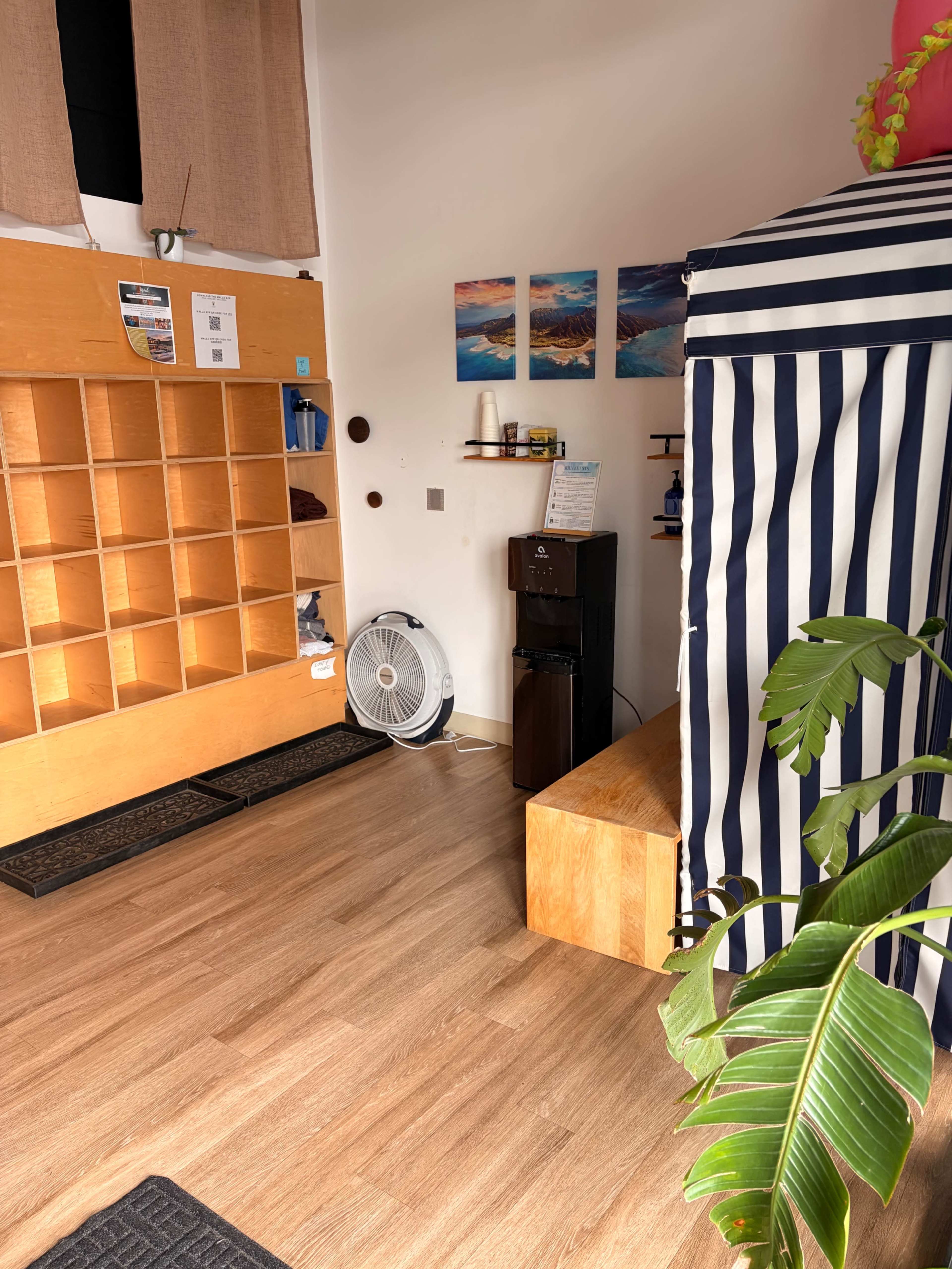 The image shows a bright, minimalist interior space with wooden shelves, a water dispenser, and a striped partition, along with a fan and a potted plant.