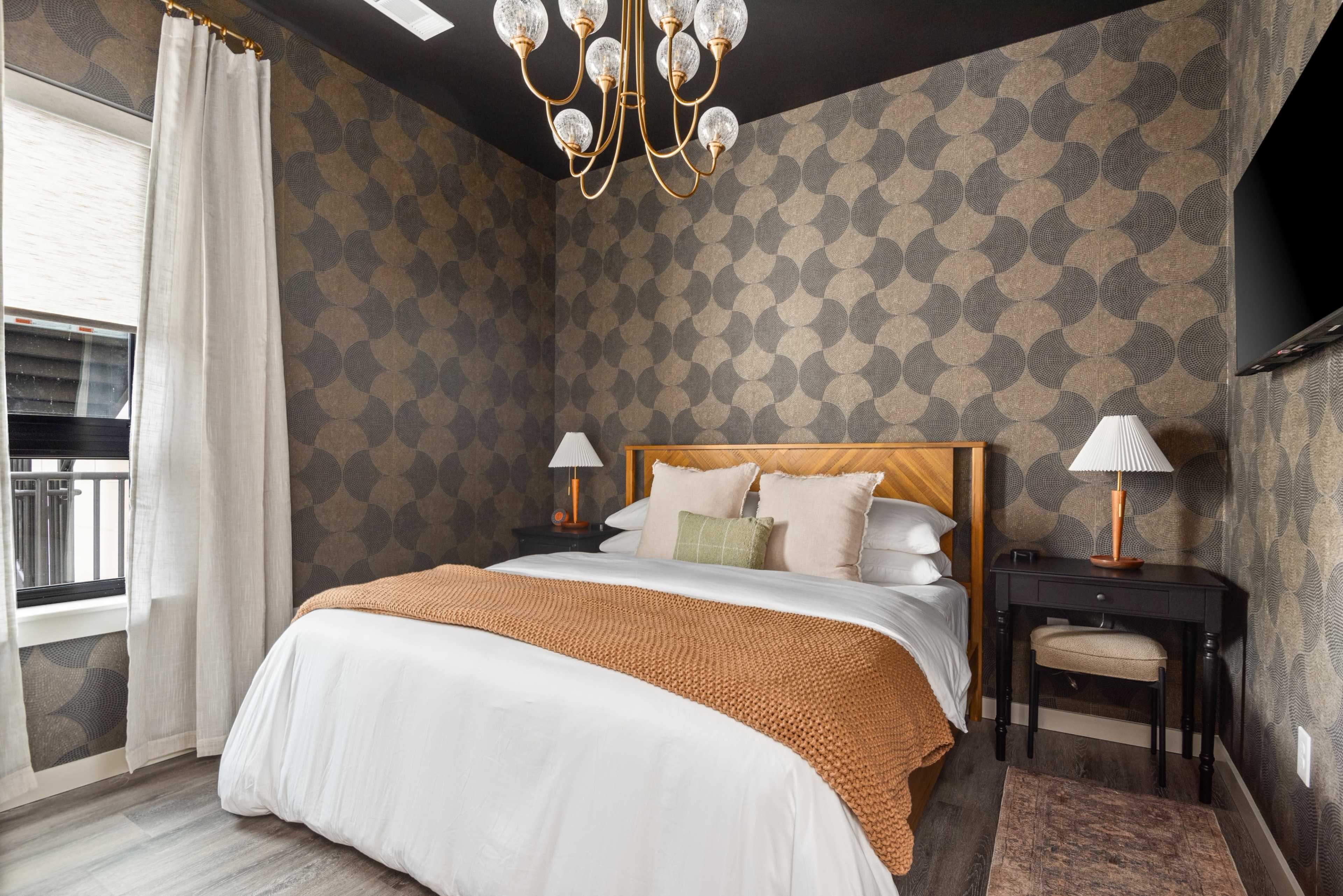 The image shows a bedroom with a patterned wallpaper, a large bed covered by a white duvet and an orange throw, lamps on either side, and a chandelier hanging from the ceiling.