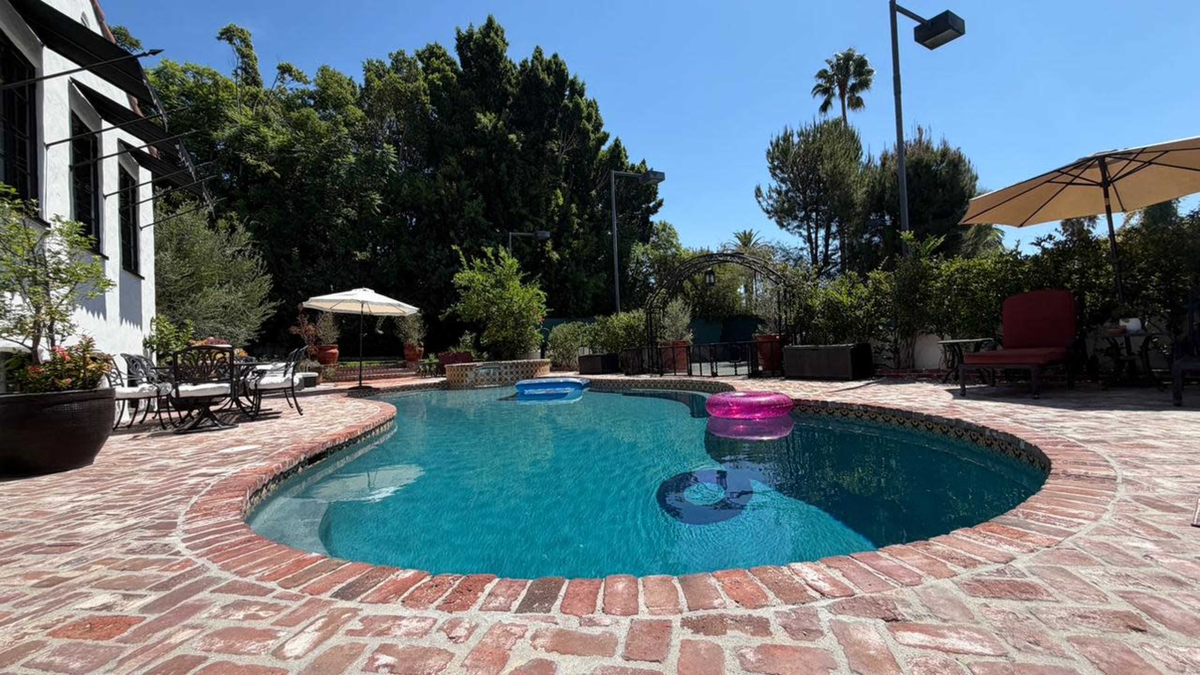 The image shows a backyard pool surrounded by brick paths, with floaties in the water and lounge chairs nearby under an umbrella.